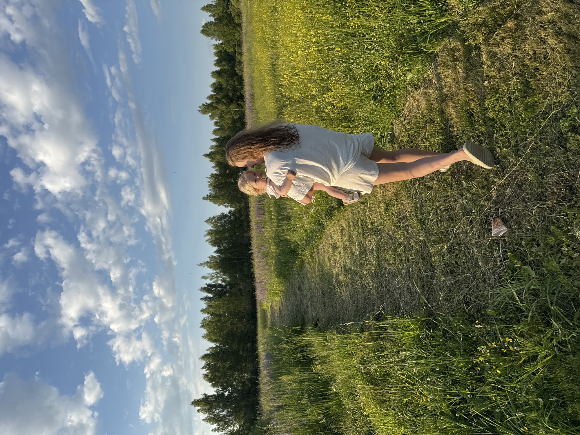 A woman carries a young child through a grassy field in the village of Koloksha, Russia, on a clear, cloudy day. The woman, facing away from the camera, wears a light-colored outfit consisting of a t-shirt and shorts. She cradles the child, who is also in light-colored clothing and has a pacifier in their mouth. A mown path runs through the field, bordered by tall green grass and scattered yellow and purple wildflowers. In the distance, a dense line of trees forms a horizon, with the bright blue sky and scattered white clouds above. The image captures a serene moment of a mother and child enjoying a walk in nature.