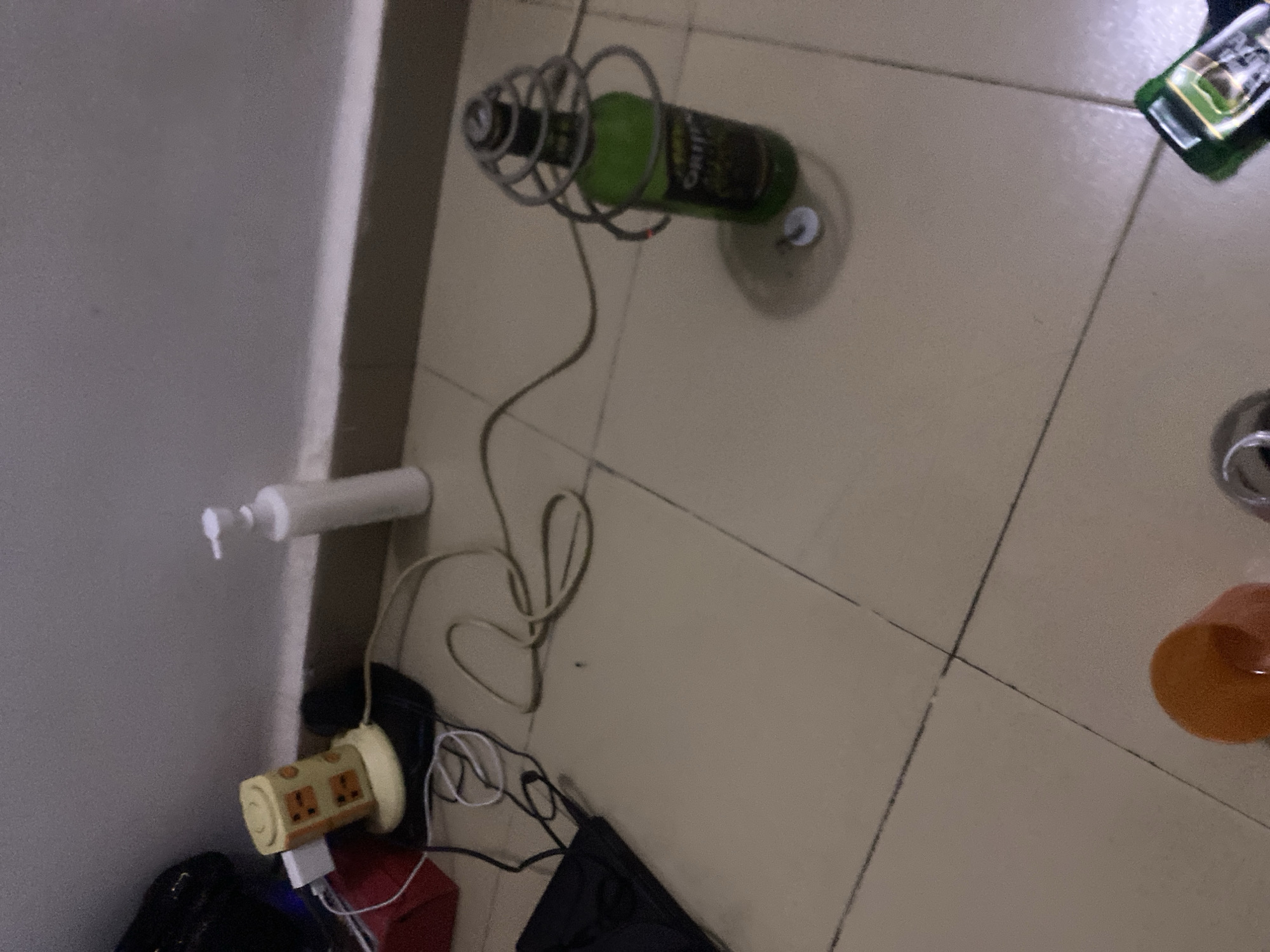 The image depicts an indoor scene on a tiled floor, likely a residential setting in Howayi, Nigeria. In the foreground, a green glass bottle with a wire coil around its neck is positioned on the floor. Adjacent to the bottle, a small silver cap is visible. To the left, a yellow cylindrical object with a face-like design and electrical plugs is connected to a power strip. A white lotion dispenser stands upright further to the left. A black object, possibly a bag or piece of electronics, lies on the floor. The overall environment suggests a temporary setup or a collection of miscellaneous items in a room.