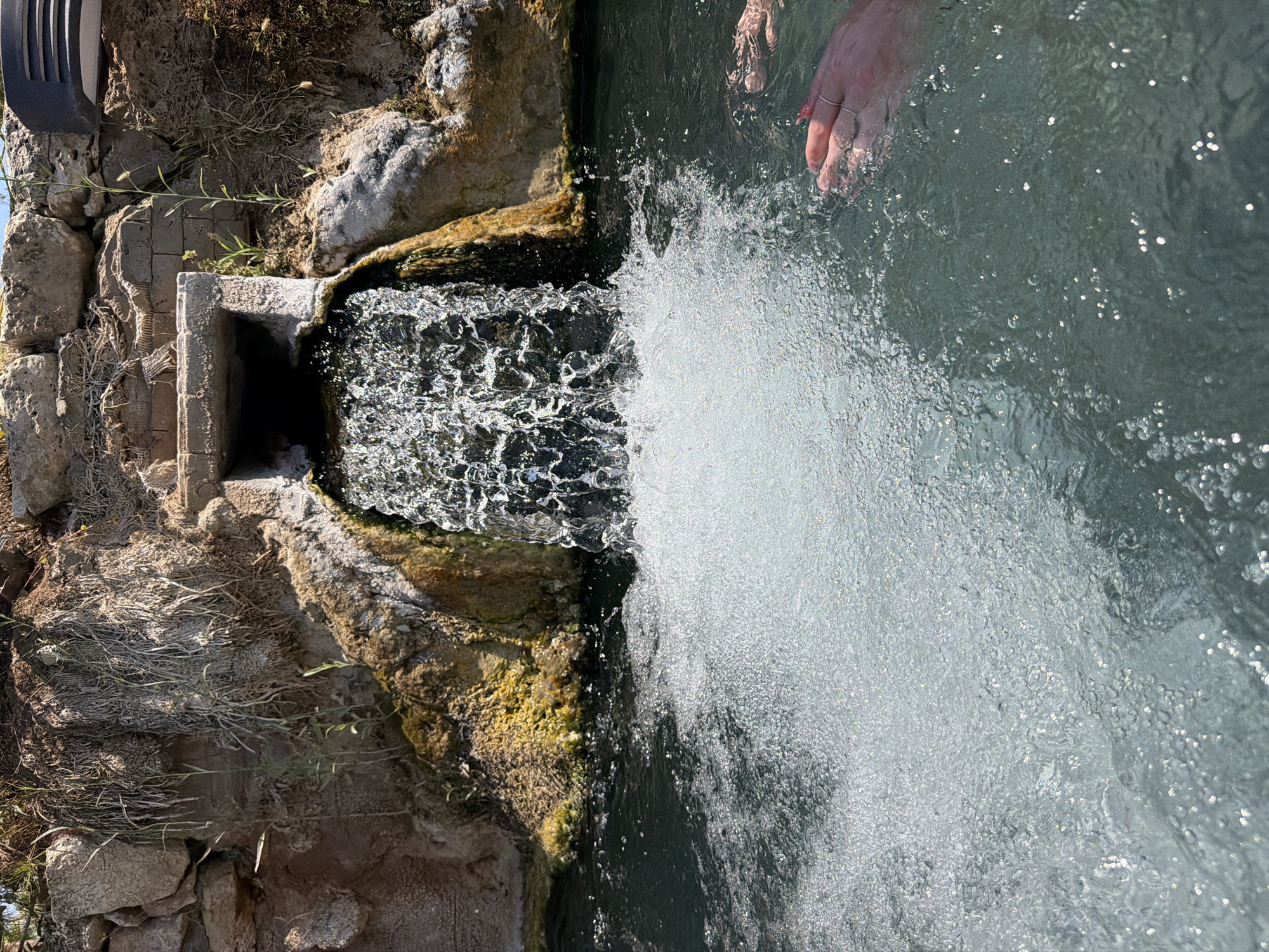 An overhead view captures a natural hot spring waterfall cascading into a pool of turquoise water. The source of the waterfall is a concrete pipe embedded in a rocky, moss-covered embankment. The water is turbulent and bubbly, indicating aeration from the falling water. Two individuals' hands are visible submerged in the water, one on the right side of the frame and another near the center, their fingers splayed. The overall scene is one of relaxation and natural beauty, likely a tourist destination. The text states the location is Pamukkale, Turkey, known for its mineral-rich thermal waters and white travertine terraces.