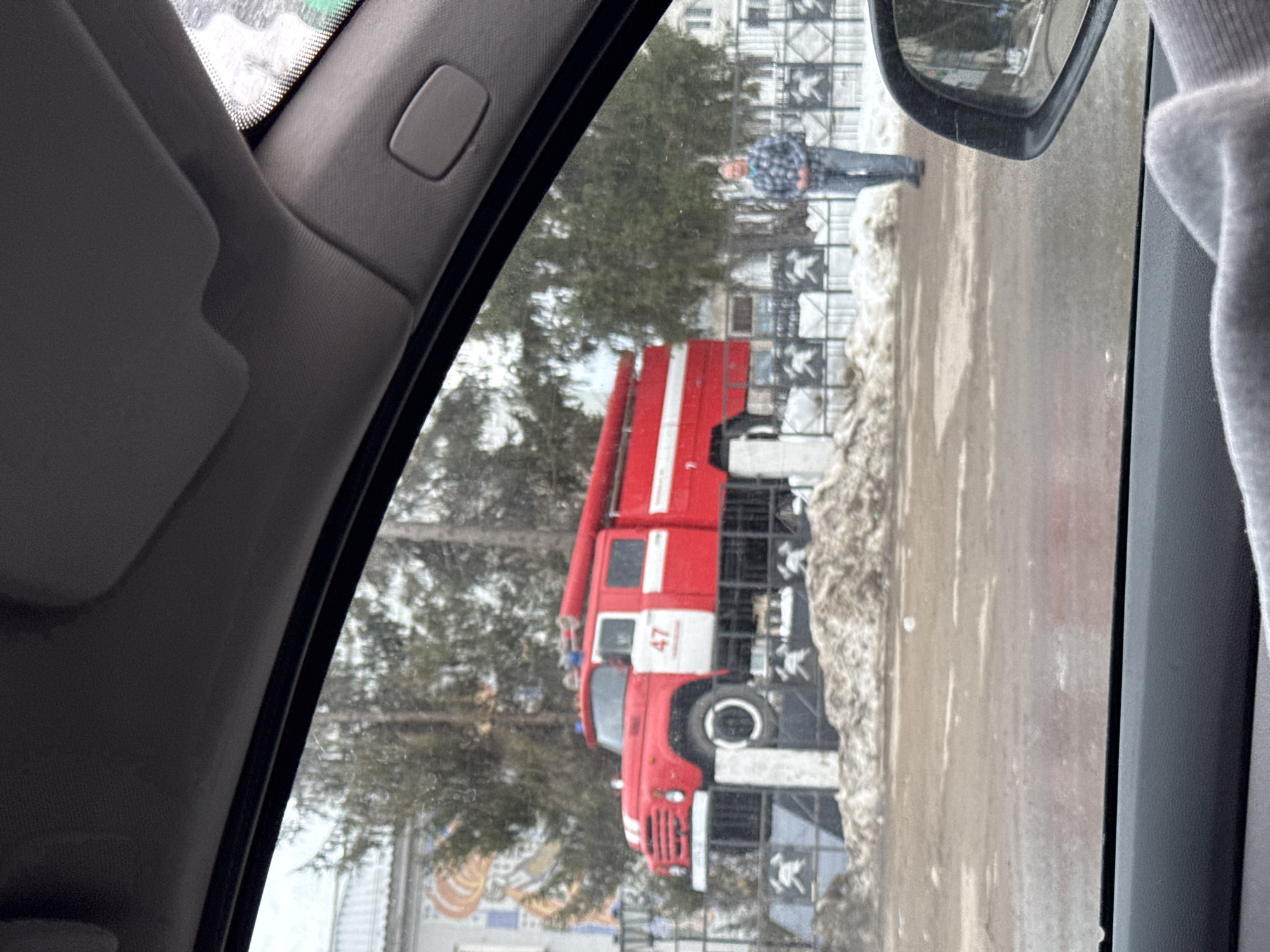 The photograph depicts a red fire engine truck parked on a street in the city of Chaikovsky, Russia, as seen from inside a car. The vehicle is parked on the side of a road with some melting snow and a fence with decorative elements. A person in a plaid shirt is visible in the background, standing by the fence. The view is framed by the car's interior, including the sun visor and side mirror. The environment appears to be a cold, overcast day.