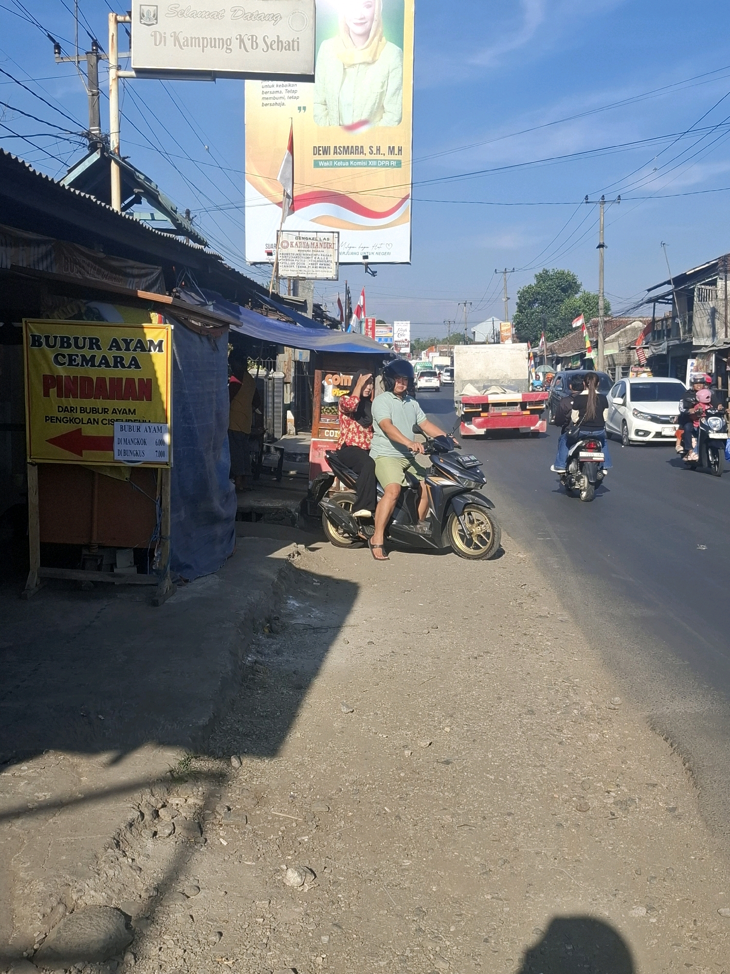 The image captures a street scene in Lembursawah, Indonesia, on a sunny day. A prominent yellow sign advertising "BUBUR AYAM CEMARA Pindhahan" (Chicken Porridge Cemara Relocation) is affixed to a wooden stand on the left. The sign indicates that the porridge is from "DARU BUBUR AYAM PENGOLAN CISULDEULL" and lists prices for "Di Mangkok" (in a bowl) at 6,000 and "Di Bungkus" (packaged) at 7,000. A red arrow points to the left, indicating the direction of the relocated shop.
In the foreground, a man wearing a light blue polo shirt and light green shorts rides a black motorcycle. He is looking forward and appears to be navigating the street. Behind him, a woman with long dark hair is a passenger on the same motorcycle. Further down the street, a white car is visible, and on the right side of the road, another motorcycle carries a woman and a child.
Various structures line the street, including stalls with blue awnings, buildings with corrugated metal roofs, and utility poles with a tangle of wires. A large billboard featuring a woman in a yellow headscarf and the text "SELAMAT DATANG DI KAMPUNG KB SEHATI" (Welcome to Healthy KB Village) is visible in the background. Indonesian flags are also present, attached to some of the buildings. The road surface is a mix of asphalt and dirt, with some gravel and debris. The overall atmosphere suggests a busy, active urban environment.