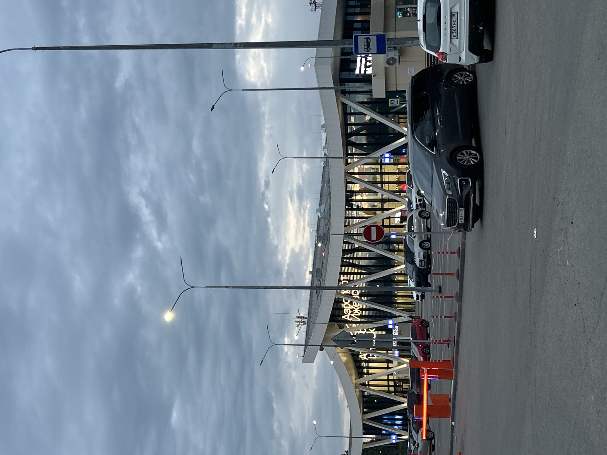 The image shows the exterior of the Izhevsk Airport (АЭРОПОРТ ИЖЕВСК) in Izhevsk, Russia. The airport building has a modern architectural design with a curved roof and a glass facade. The sky is overcast, and streetlights are illuminated. Several cars are parked in the foreground, and a retractable barrier is visible on the left side of the image. A bus stop sign is present on the right. The scene appears to be a parking lot or drop-off area outside the airport terminal.
