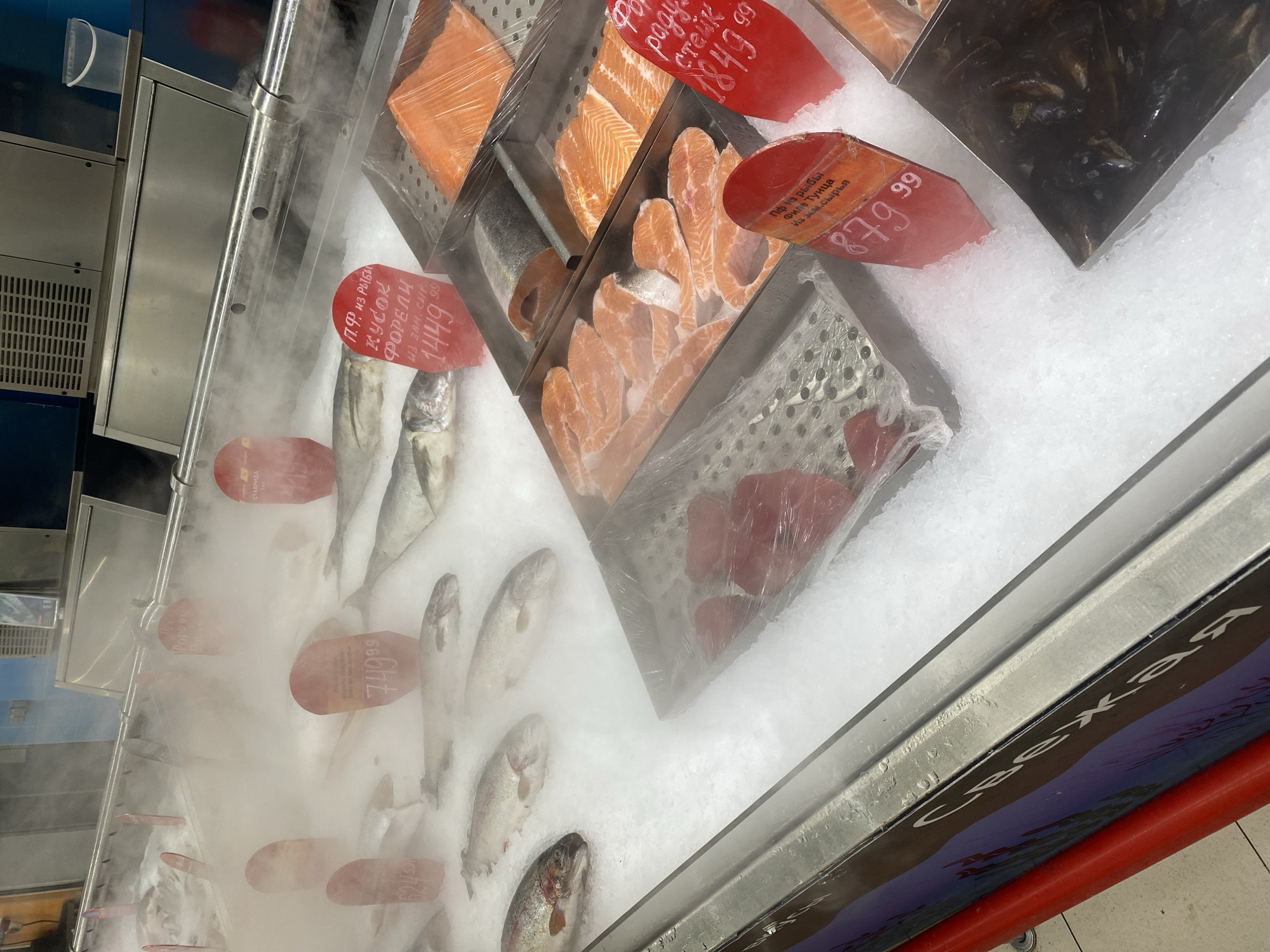 The image shows a refrigerated display case at a seafood counter, likely in a supermarket. The case is filled with crushed ice to keep the fish fresh. Several types of fish are displayed, including whole fish (likely mackerel or similar), salmon fillets, and what appear to be tuna steaks. Some shellfish (possibly mussels) are also present. Each item has a price tag stuck into the ice. The price tags are predominantly red and display prices in what appears to be Russian currency (rubles). The display case is generating a mist, which obscures some of the fish. The words "свежая" (fresh) are visible on the front of the display case. The image has a slightly angled perspective.