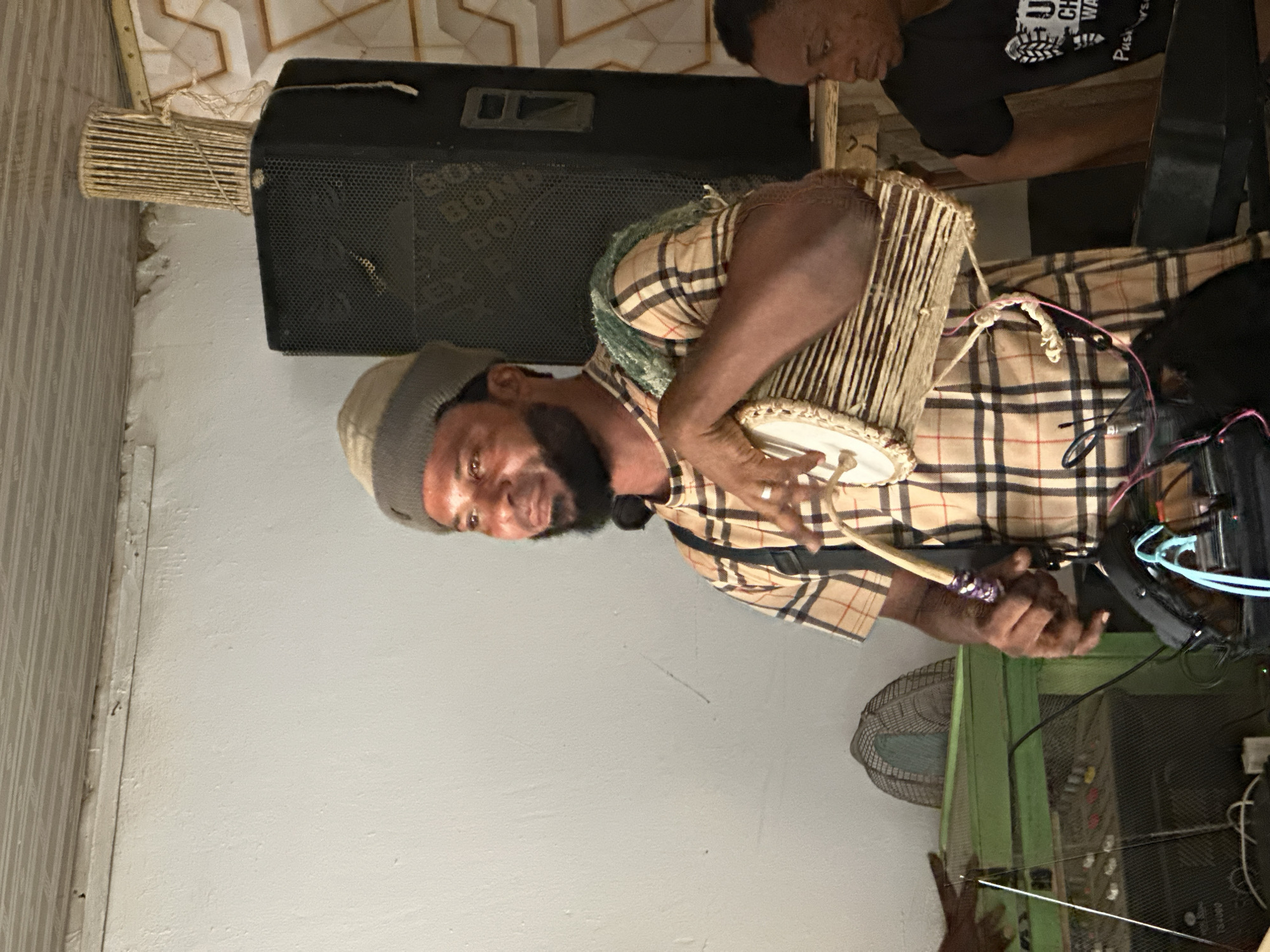 The image captures an indoor musical performance or rehearsal, featuring two men in a candid, upward-angled shot. The main subject, positioned centrally, is a man with dark skin, a neatly trimmed beard, and a striped beanie in shades of gray and beige. He wears a distinctive short-sleeved plaid shirt, resembling a Burberry check, and is actively playing a drum. This drum, which appears to be a small hand drum or a style akin to a talking drum, is strapped across his body. His left hand rests on the drumhead while his right hand holds a slender drumstick, ready to strike. He looks directly at the camera with a slight, engaging smile, conveying enthusiasm.
In the background, to the right, a second man with dark skin is partially visible. He is wearing a black t-shirt, on which faint, incomplete white text such as "UN," "CH," and "WA" can be discerned. He appears to be observing or also involved in the musical activity.
The setting includes various pieces of musical equipment. Behind the main drummer is a large, black speaker with a perforated grill, on which some indistinct text is barely visible. A unique, tall, cylindrical percussion instrument, possibly a shaker or rainstick made of numerous parallel wooden tubes, stands vertically next to the speaker. To the right, an assortment of electronic devices, cables, and possibly an audio mixer are visible. A circular fan with light blue blades is partially obscured behind these electronics. The room features plain white walls and a light-colored ceiling with a decorative geometric pattern. The overall ambiance suggests a casual, intimate musical session, likely in Lagos, Nigeria, given the provided context. The time of day is not evident due to the indoor artificial lighting.