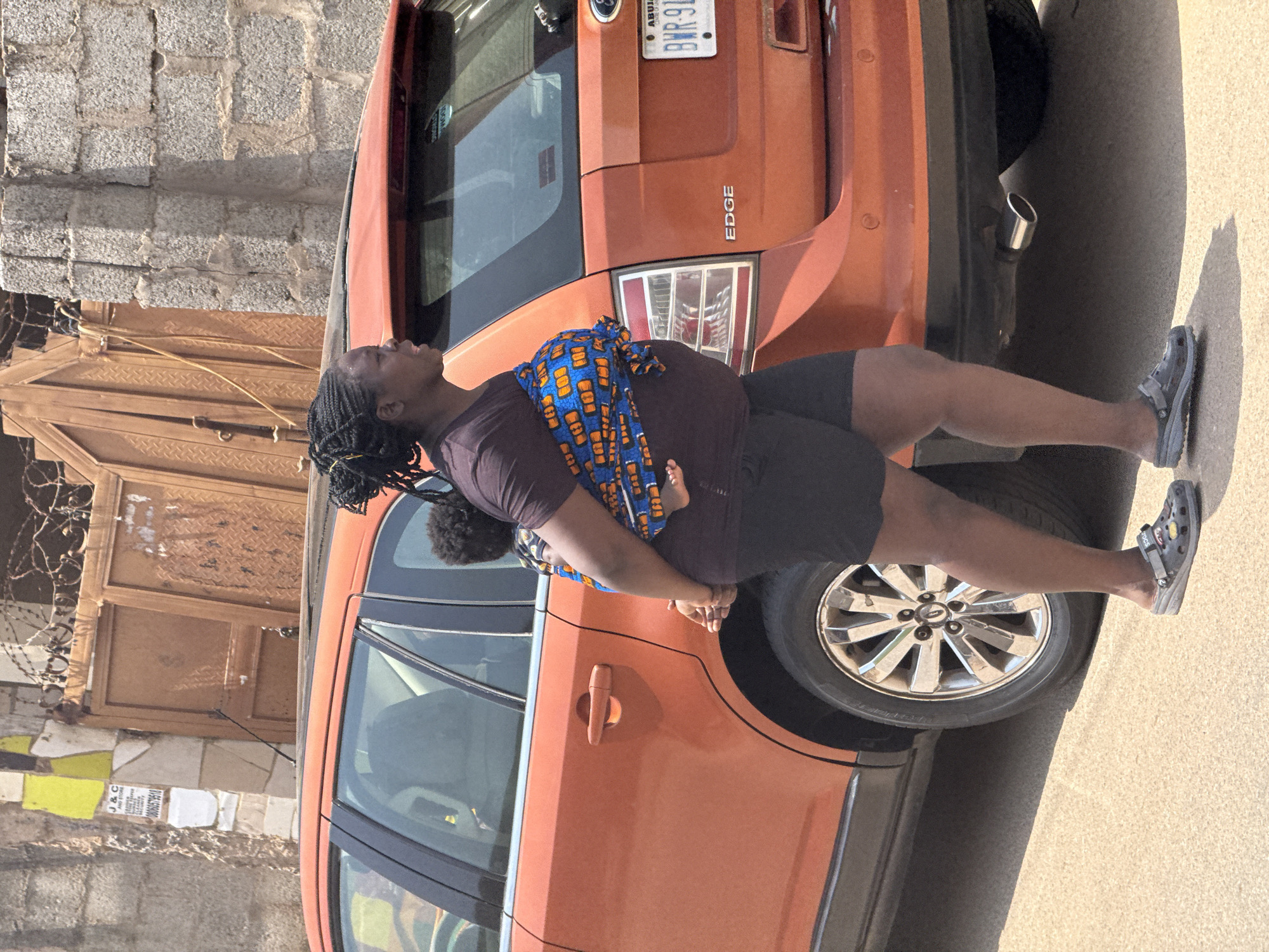An outdoor medium shot captures a Black woman carrying a baby in a sling, standing next to an orange Ford Edge SUV in Abuja, Nigeria. The woman is positioned on the right side of the frame, facing left. She has dark braided hair and is wearing a dark-colored t-shirt and black shorts. The baby, also Black, is wrapped in a blue and orange patterned fabric, with only the baby's head and legs visible. The Ford Edge is a vibrant orange color and takes up a significant portion of the background. The license plate reads "ABUJA BWR 91M". The scene appears to be on a street, with a textured concrete or asphalt ground. Behind the car and woman, a brick wall and a large brown metal gate with intricate patterns are visible. The gate is partially obscured by barbed wire. The overall lighting suggests a sunny day.