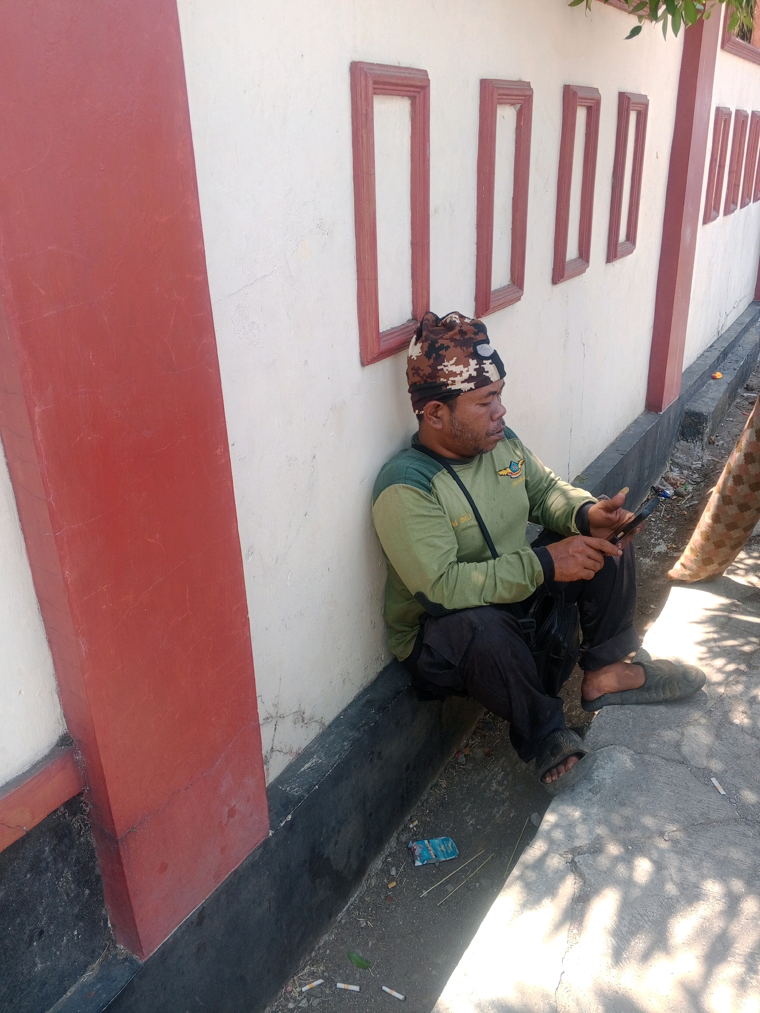A man is sitting against a white wall with red trim in Giritirto, Indonesia, looking at his phone. He is wearing a camouflage head covering, a green long-sleeved shirt with a patch on the left chest, black pants, and dark-colored slip-on sandals. A black bag is slung over his shoulder. The wall has several rectangular frames on it. In front of the wall is a dark grey concrete base. The ground in front of the base is made of concrete with some dirt and debris, including cigarette butts and a small blue and white packet. Sunlight casts dappled shadows on the ground. To the right of the man, another person's leg wearing camouflage pants and a beige sandal is visible, suggesting another individual is present but partially out of frame. A tree branch with green leaves is visible at the top right of the image.