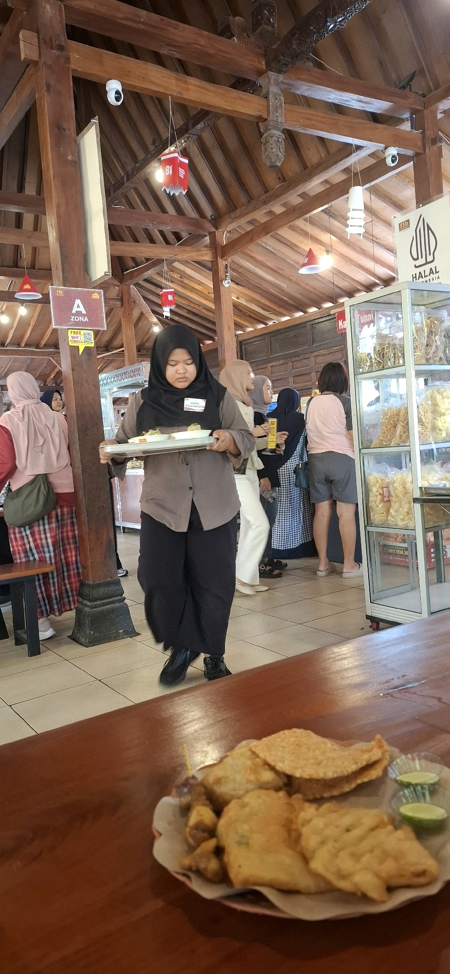 In Colomadu, Indonesia, a woman in a black hijab and brown uniform is carrying a tray of food in a restaurant. She is walking past a display case filled with snacks. The restaurant has a rustic wooden interior with exposed beams and a tiled floor. Other patrons are visible in the background, some standing in line at the display case. A plate of fried snacks and lime wedges is in the foreground on a wooden table.