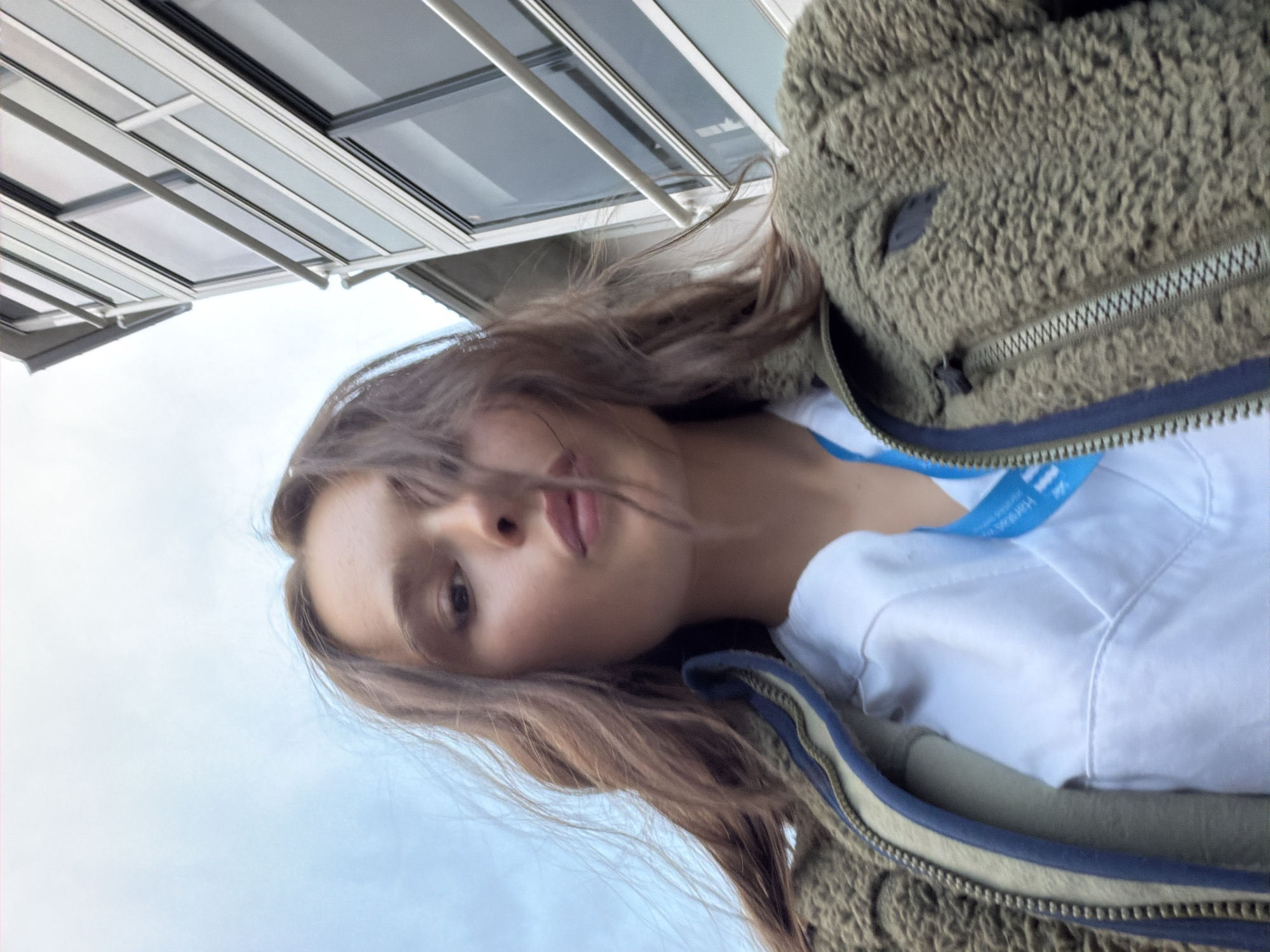 A young woman with long brown hair, wearing a white shirt and a fuzzy olive-green jacket, poses for a selfie outdoors. She is looking into the camera with her mouth slightly parted. Behind her, a modern building with large windows and horizontal railings is visible, along with a cloudy sky. A blue lanyard with white text is partially visible around her neck, suggesting she may be associated with the city of Harstad, Norway.