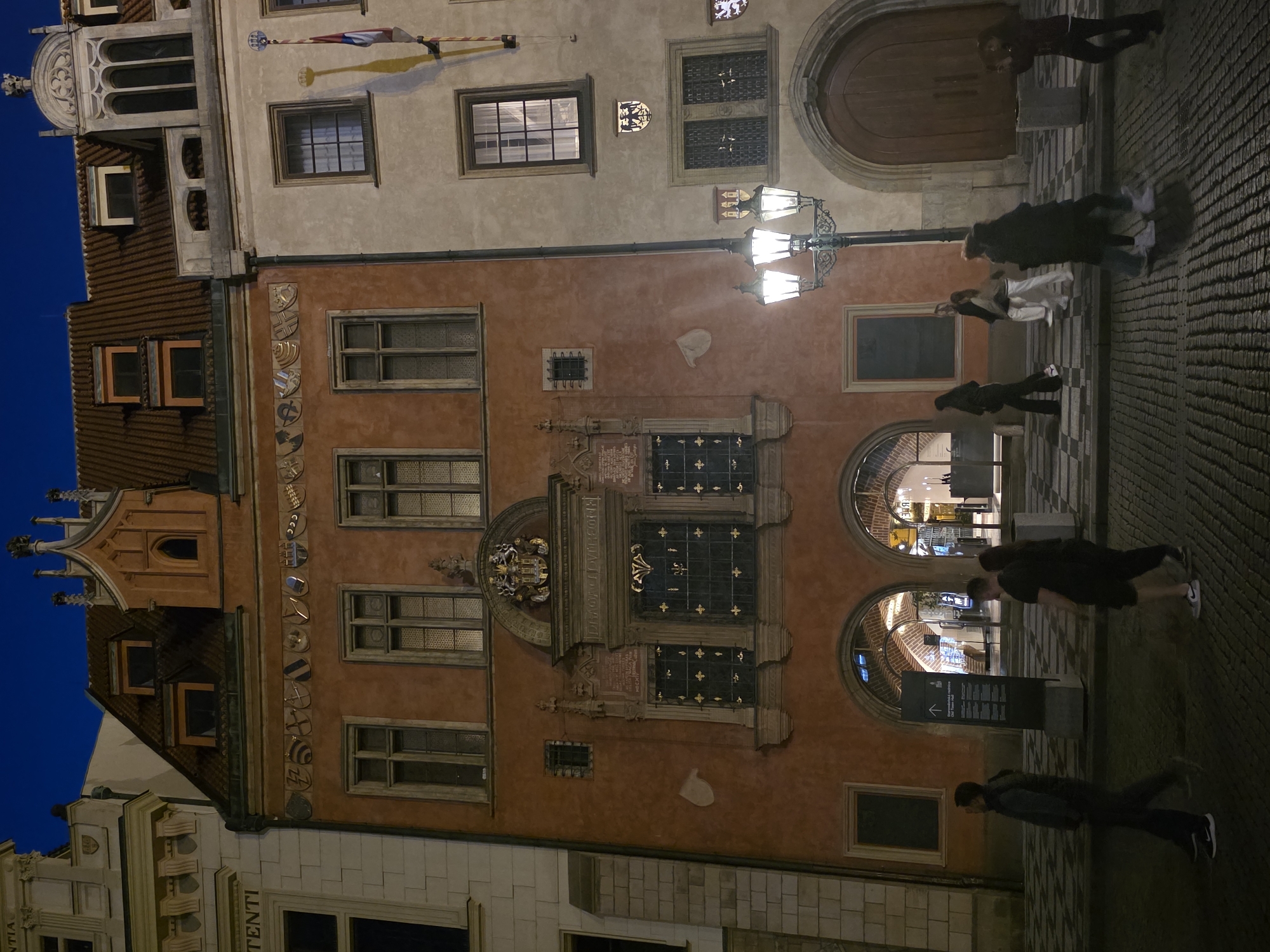 The image depicts a street scene at night in an unknown city. The main subjects are two historic buildings. The building on the left is reddish-orange with ornate architectural details, including multiple windows, decorative shields above the windows, and a crest above a central window. The building on the right is lighter in color and features a large arched doorway and several windows. A lamppost with three illuminated globes stands in the foreground, casting light on the cobblestone street. Several people are walking on the street, some in groups and others individually, appearing to be tourists or pedestrians. The sky is dark blue, indicating it is nighttime. The scene is illuminated by artificial lights from the buildings and the lamppost, creating shadows and highlighting the architectural features.