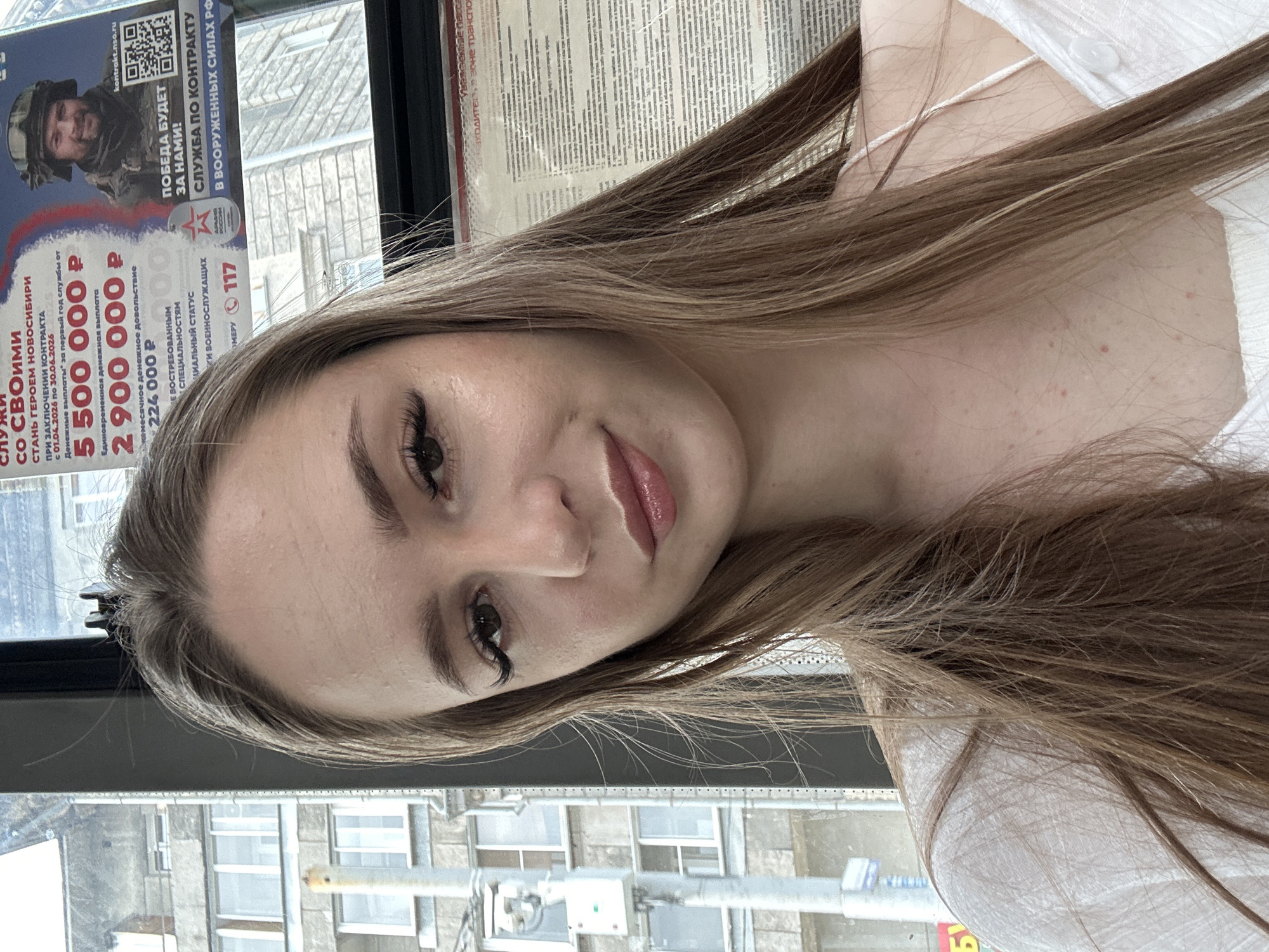 In this portrait, a young Caucasian woman with long, light brown hair is positioned in the foreground, her face slightly turned towards the camera. She is looking directly at the viewer, her expression neutral. Her makeup includes noticeable eyeliner and mascara, enhancing her eyes, and a glossy lip product. Her skin appears clear, with a few small blemishes visible on her décolletage. She is wearing a white top with thin straps.
Behind her, through a window, a city landscape is visible. The background shows a multi-story building with numerous windows, suggesting an urban setting. A poster is also partially visible, featuring Russian text and an image of a soldier. The lighting suggests an overcast day, with soft, diffused light. The overall impression is that of a candid selfie taken indoors or in transit, possibly on a bus or train, given the presence of text and the window. The specific location in the city is unknown.