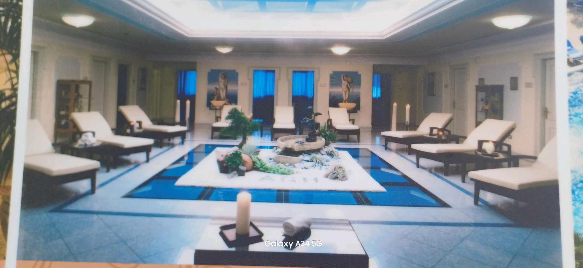 The image depicts a luxurious indoor spa or relaxation area, possibly located in Abano Terme, Italy, a city known for its thermal baths. The room features a tranquil and opulent design. In the center, a shallow pool or water feature with white decorative stones and a small bonsai tree creates a focal point. Around this central area, several recliners with white cushions are strategically placed, suggesting a space for guests to relax and unwind. The floor is tiled with a pattern of blue and white squares. In the background, there are tall blue curtains covering windows or doorways, and decorative artworks depicting figures. Lighting appears to be provided by recessed ceiling lights and perhaps ambient lighting from the artworks and candles. The overall impression is one of serenity and upscale comfort.