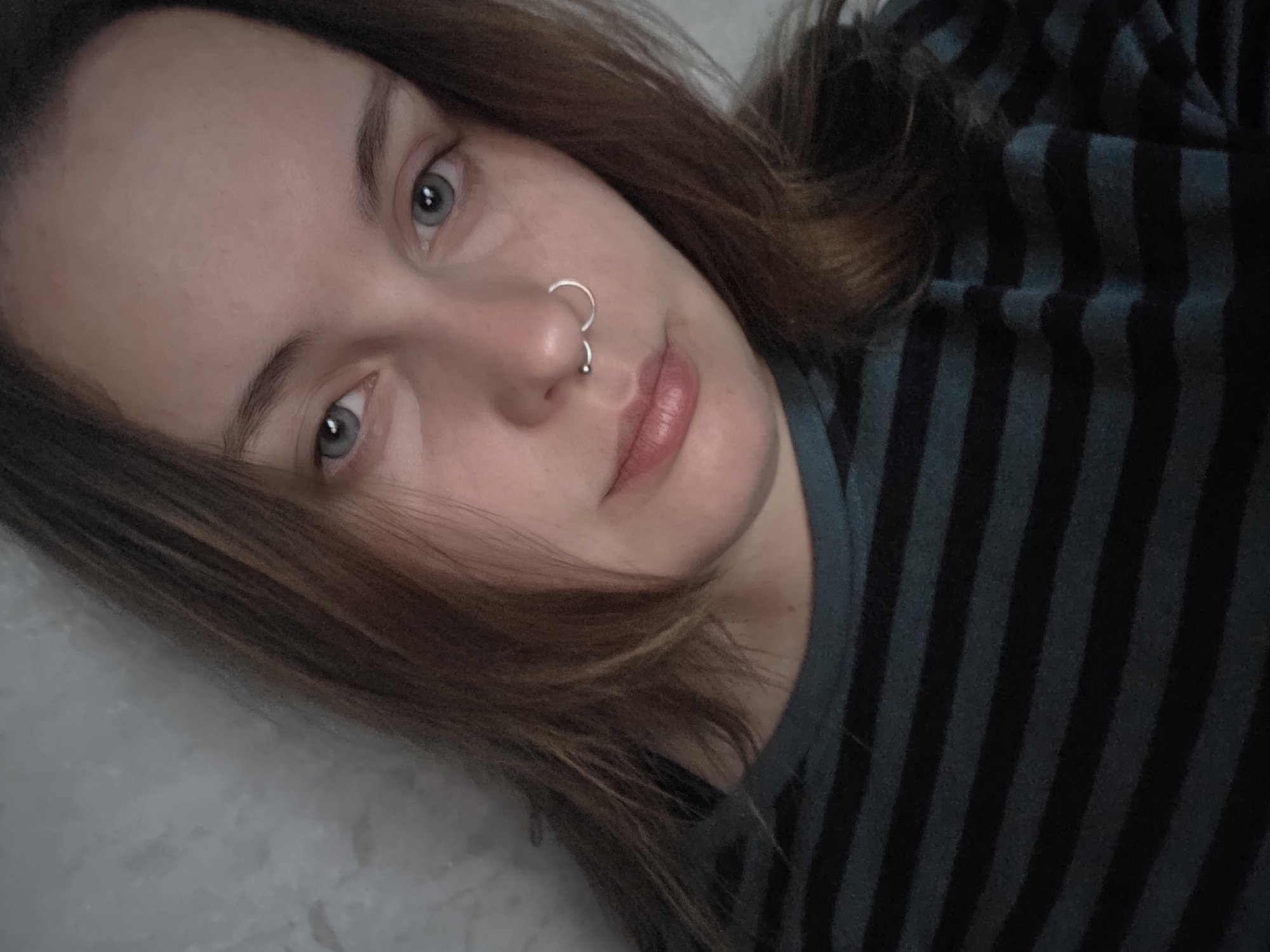 A close-up, head-and-shoulders selfie of a young Caucasian woman with light brown hair and blue eyes. She is wearing a dark green and black striped long-sleeved shirt. She has a septum piercing. The background appears to be a textured gray wall. The image is undated and its location is unknown.