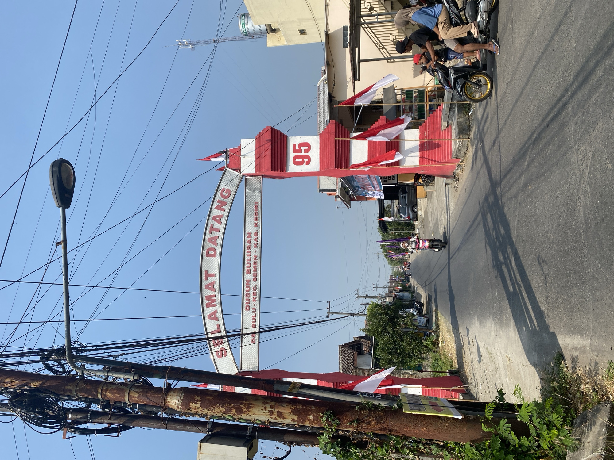A street scene in the city of Kediri, Indonesia, is captured under a clear blue sky. A large red and white archway, a prominent landmark, spans the road, bearing the inscription "SELAMAT DATANG" (Welcome) in red lettering. Below this, smaller text reads "DUSUN BULUSAN DS. BULU KEC. SEMEN KAB. KEDIRI". The number "95" is prominently displayed on the archway's pillar. Indonesian flags are strategically placed on the archway and along the street, suggesting a festive or patriotic occasion.
The road is paved and appears to be a main thoroughfare, with several motorcycles visible. In the foreground, a family of three, including a child, is seated on a black motorcycle. The child is wearing a red cap. Other individuals are also present on motorcycles further down the street. To the left of the road, a weathered utility pole is heavily laden with electrical wires, a common sight in urban Indonesian landscapes. On the right side of the road, buildings and smaller shops are visible, with a water tank on top of one structure. The environment suggests a typical urban or semi-urban setting in Indonesia.