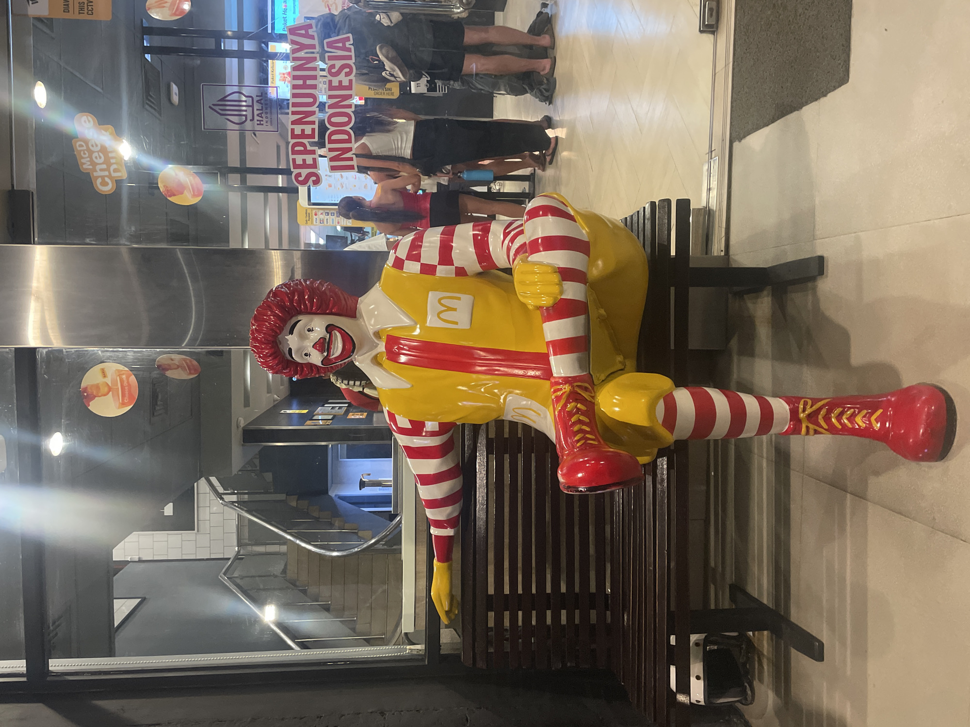 The photograph shows a statue of Ronald McDonald sitting on a bench outside a McDonald's restaurant in Denpasar, Indonesia. The statue is wearing Ronald McDonald's signature yellow jumpsuit with the McDonald's logo, red and white striped leggings, and red shoes. The restaurant's exterior is visible through a glass wall and shows customers standing in line.