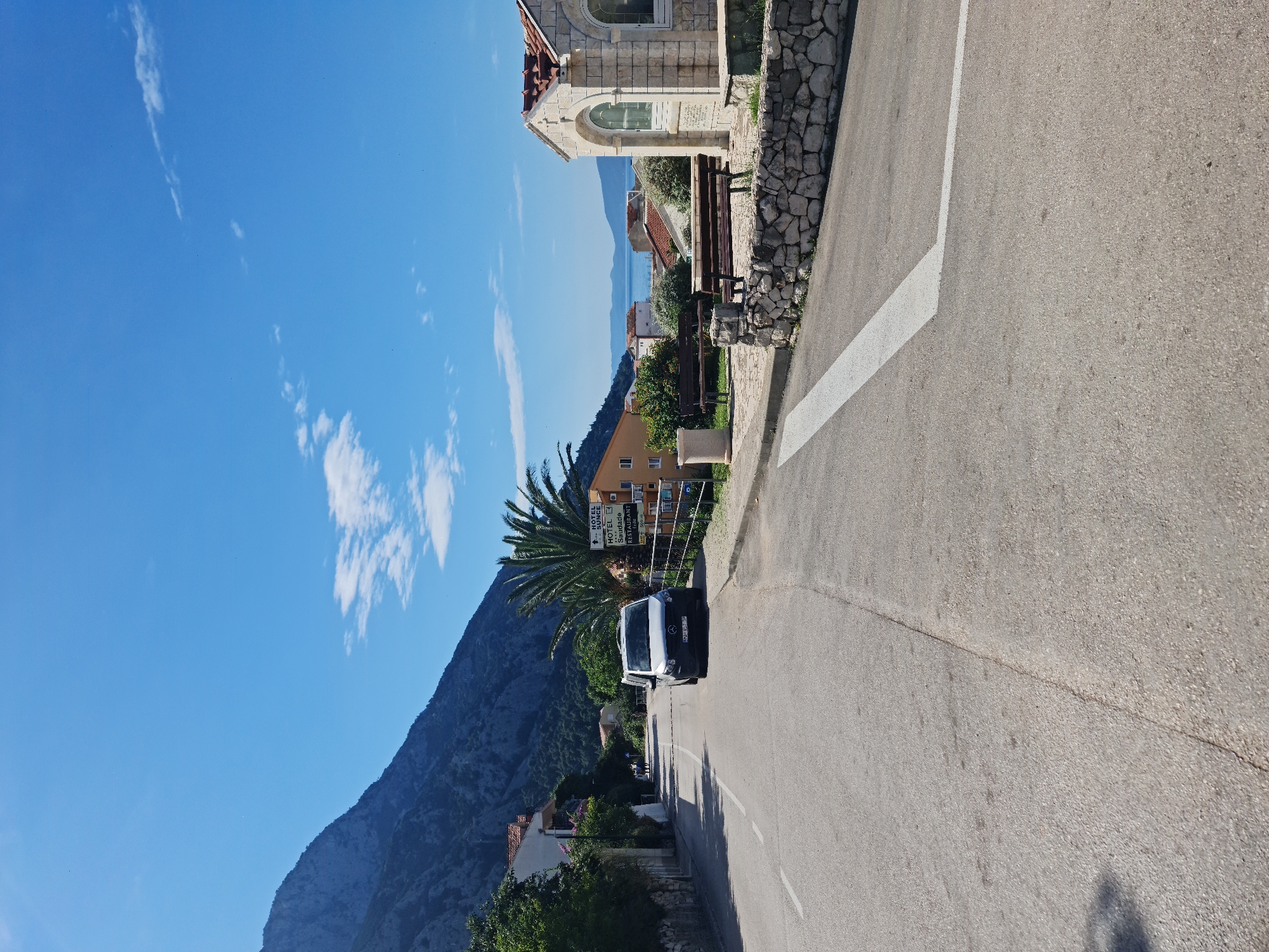 The image depicts a street scene in Općina Gradac, Croatia. A grey van is parked on the right side of the asphalt road. To the right of the van, a signpost indicates "HOTEL SUNCE" and "HOTEL SAUDADE RESTAURANT". Beyond the signpost, there is a yellow building and a palm tree. Further to the right, a small stone building with an arched window is visible, with a stone wall and wooden benches in front of it. The background shows a body of water, likely the Adriatic Sea, with distant hills. A large, dark mountain dominates the left side of the frame, covered in sparse vegetation. The sky is clear blue with a few scattered white clouds. The road has a white dashed line marking the lane. There are no people visible in close proximity to the camera, though two figures appear to be walking in the distance on the left side of the road. The overall scene suggests a warm, sunny day in a coastal town.