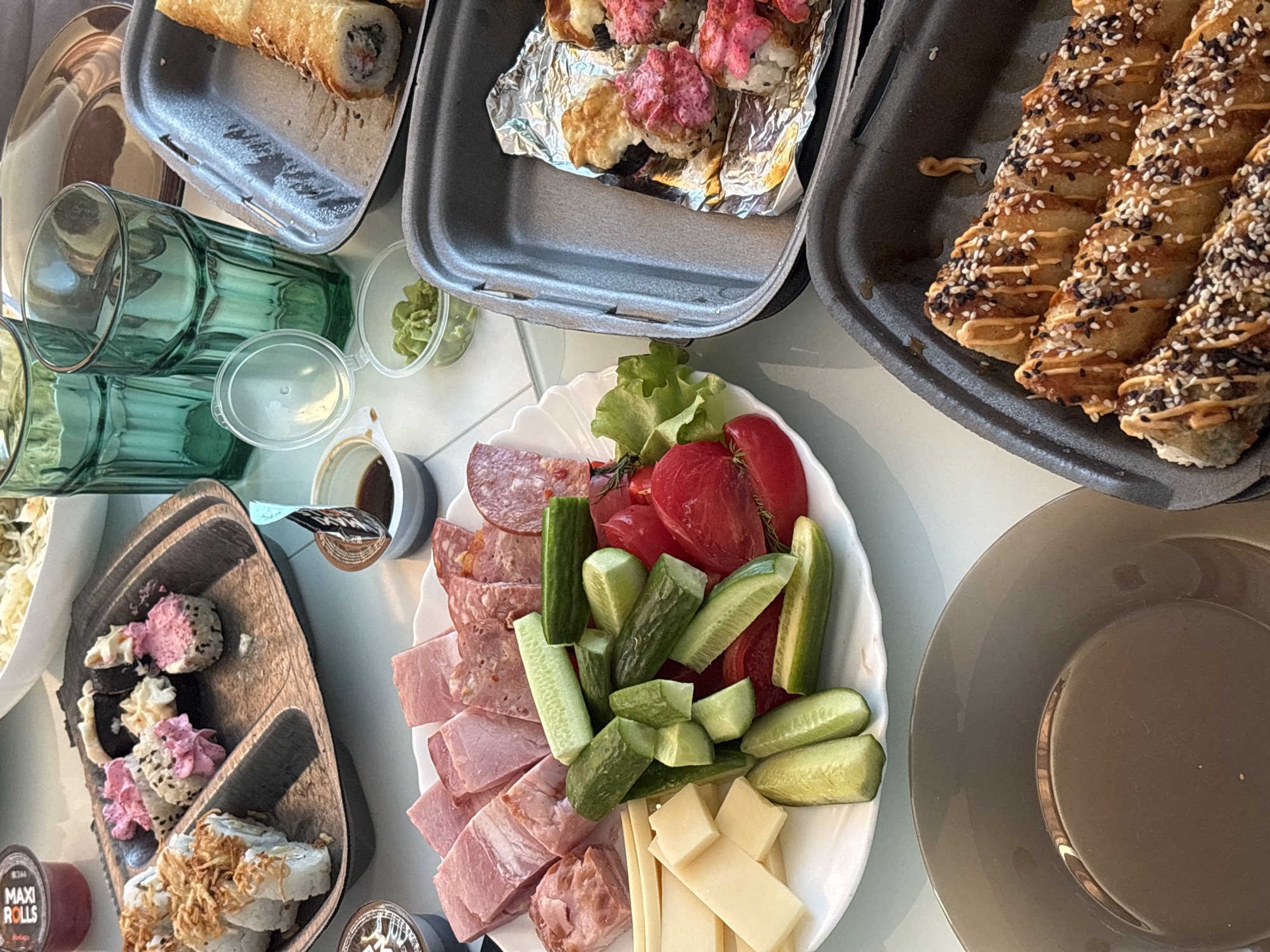 The image shows a table laden with a variety of food, suggestive of a meal shared by multiple people. Various containers hold sushi rolls: one with fried rolls, another with rolls topped with pink sauce, and a third with rolls drizzled with sauce and sesame seeds. A wooden platter contains a different type of sushi rolls. The center of the table features a white plate with a selection of meats, cheeses, sliced cucumbers, and tomatoes. There are also transparent glasses and condiment containers scattered around the table. A large, grey plate sits empty on the right side of the image. The scene appears to be indoors. There is no definitive evidence to confirm the location in Prikamskii, Russia.