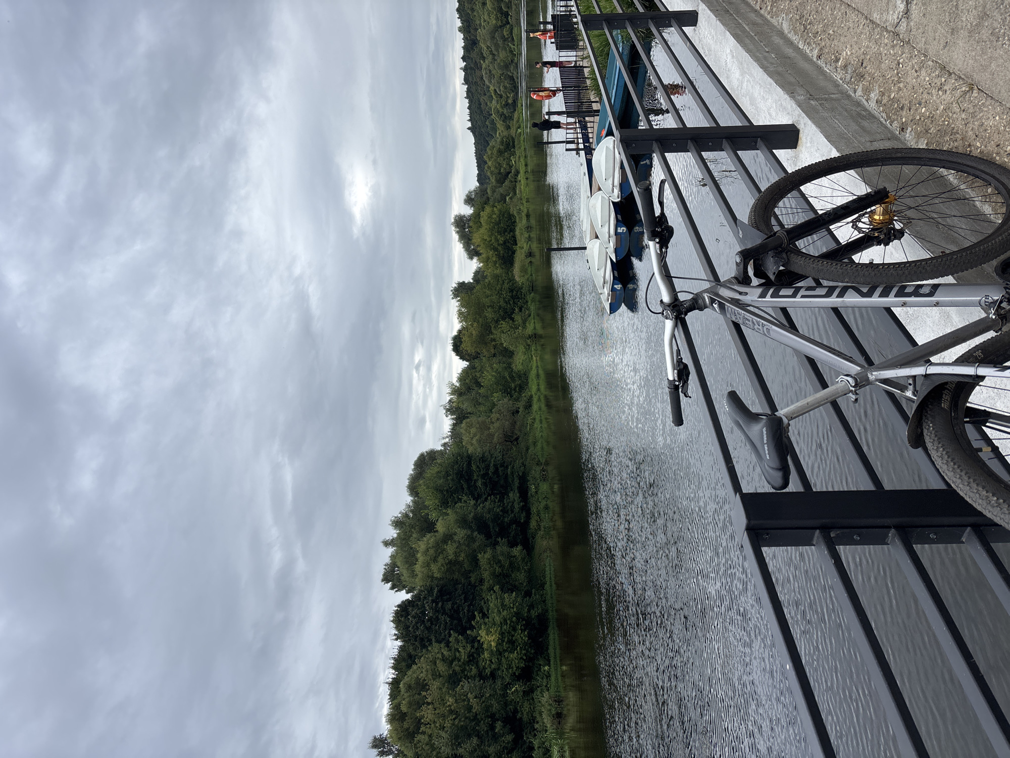 This image depicts a scene along the Moskva River in Zvenigorod, Russia, on an overcast day. In the foreground, a silver mountain bike is parked next to a black metal railing that separates a concrete walkway from the river. The bicycle's handlebars, front fork with gold accents, and partial view of its front tire and frame are visible. The river, appearing dark and rippled, flows to the left, with its surface reflecting the cloudy sky. Across the river, a dense, lush green forest lines the bank, with trees of various heights and shades.
Further down the river, a wooden pier extends into the water, with several white rowboats tied to it. Some of the boats are numbered, such as boats with the numbers 4, 5, and 8. Two individuals, dressed casually, are standing on the pier, one appearing to be interacting with a life preserver. The pier and boats are reflected in the water. The background beyond the river and forest shows a rolling landscape covered in trees, suggesting a natural, somewhat rural setting within the vicinity of Zvenigorod. The sky is a uniform gray, indicating cloud cover.