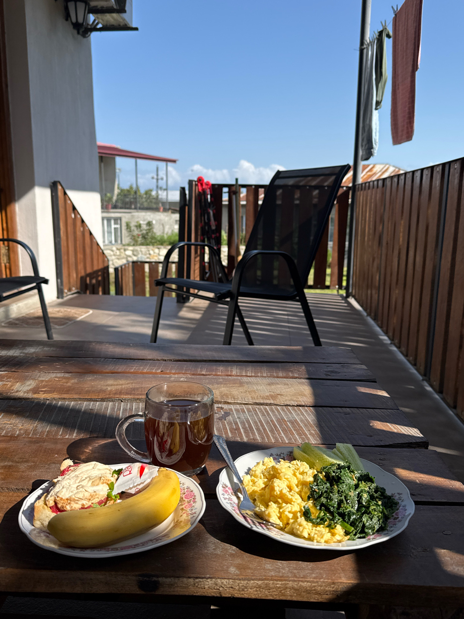 This is a close-up, eye-level shot of two plates of food and a glass of tea on a rustic wooden table. The setting appears to be an outdoor patio or balcony, with a wooden fence in the background and chairs visible. The sky is a clear blue.
On the left plate, there is a pastry with a light-colored topping and a bright yellow banana. The pastry appears to have some red filling peeking out.
On the right plate, there is a serving of scrambled eggs and a pile of dark green leafy vegetables, possibly spinach or kale. Two slices of cucumber are also visible on this plate. A fork rests on the edge of this plate.
A clear glass mug filled with a dark liquid, likely tea or coffee, sits between the two plates. The mug has a handle and is casting a shadow on the table.
In the background, there are two black outdoor chairs with metal frames and fabric seats. Beyond the chairs, a wooden fence runs horizontally, and a glimpse of residential buildings and vegetation can be seen. Clothes are hanging on a line to the right of the scene.
The image was taken in Telavi, Georgia.