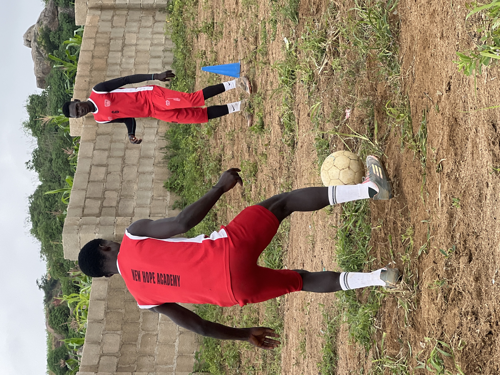 Two young men, likely teenagers, are engaged in a soccer training session on an outdoor dirt field. The man in the foreground, facing away from the camera, wears a red sleeveless jersey with "NEW HOPE ACADEMY" printed vertically in black on the back, red shorts, white socks with grey stripes, and athletic shoes. He is in the process of kicking a worn, off-white soccer ball. The second man, positioned further back and to the right, faces the camera. He wears a red jersey over black long-sleeved undershirt, red shorts, and athletic shoes. A blue training cone is visible behind him. The environment is rural, with a rough, unpaved ground covered in dry grass and some green vegetation. A low, light-colored brick wall forms a backdrop, with lush green trees and foliage, including corn stalks, growing behind it. In the distance, large rock formations are visible, suggesting a hilly terrain. The sky is overcast. The scene appears to be in a less developed area, consistent with the outskirts of Jos, Nigeria, where such informal training grounds are common.