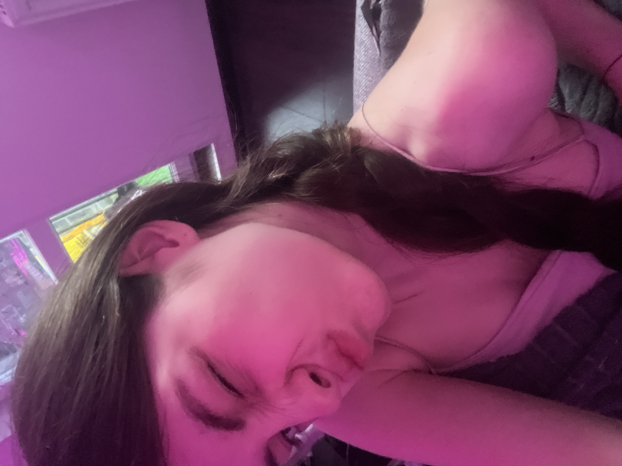 A close-up, horizontally oriented image displays a person's face and upper body under pink lighting. The person has dark hair and closed eyes. They appear to be lying down. The background includes a window with partially visible objects outside and a dark, soft surface, possibly a blanket or cushion. No information suggests the image was taken in Saint Petersburg, Russia.
