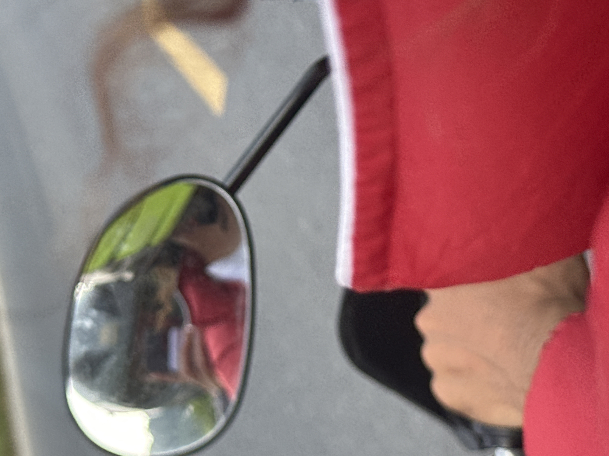 A close-up shot focuses on the left side-view mirror of a vehicle, likely a motorcycle or scooter, in Vị Xuyên Commune, Vietnam. The mirror has an oval shape with a metallic rim, and its dark stem extends diagonally towards the upper right.
To the right of the mirror, a person's arm is visible, dressed in a vibrant red garment that features a distinct vertical white stripe along its side. Below the sleeve, a light-skinned hand firmly grips a dark, possibly black, handle or control, suggesting the person is operating or about to operate the vehicle.
The surface of the mirror reflects a blurred scene: indistinct green shapes, likely foliage or greenery, are prominent, along with hints of red and darker, shadowy forms, possibly another person or the person's own reflection. The background behind the mirror and the arm is uniformly blurred in shades of gray, consistent with a road surface or asphalt. In the upper left quadrant of the image, a bright, diffused yellow shape suggests a light source or a reflected object. The scene appears to be outdoors during daytime. No text is visible in the image.