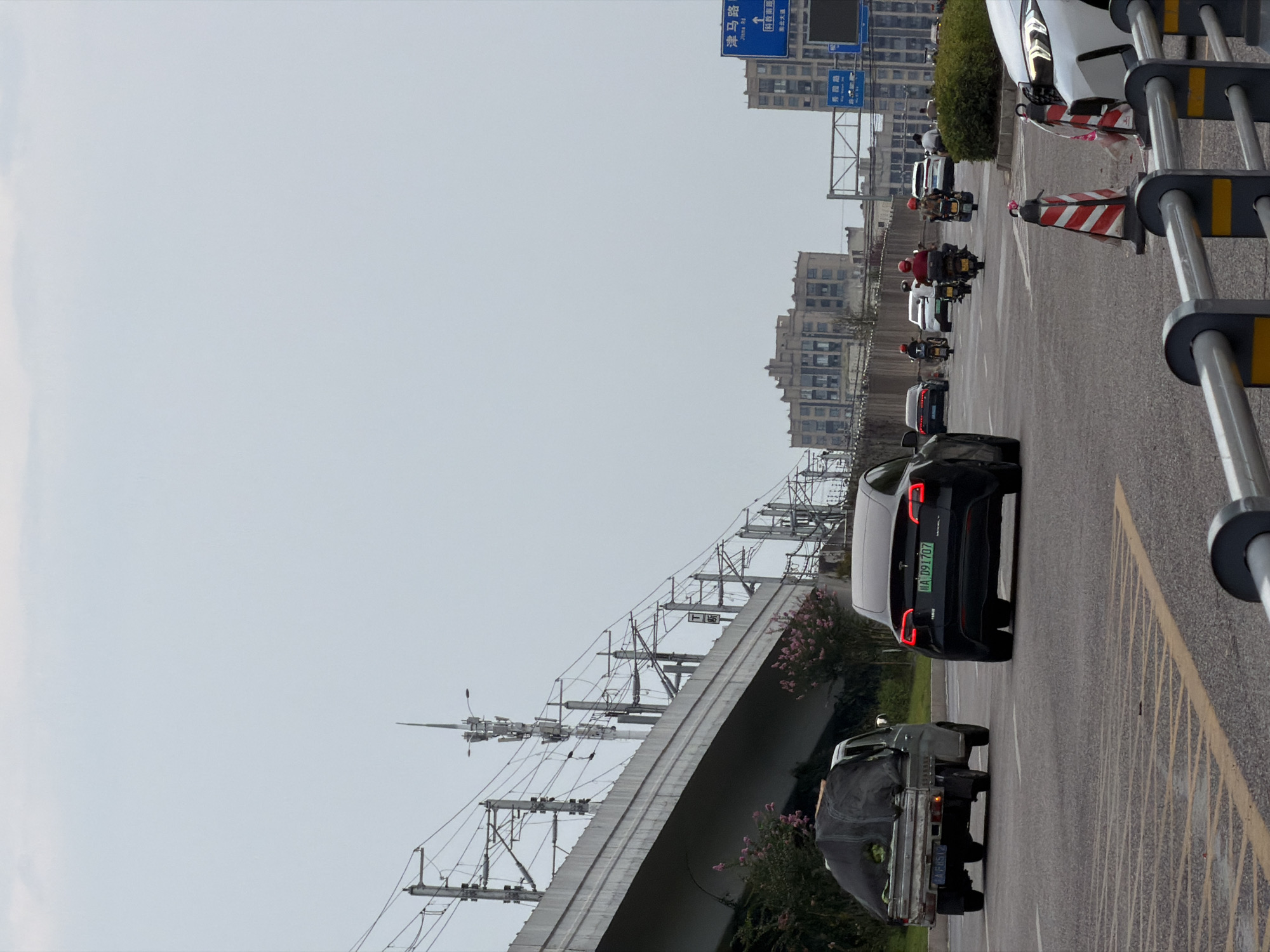 The image shows a road scene in Jiangjin, China, with an elevated railway track and overhead power lines. In the foreground, a black Tesla Model Y is driving on the road, its brake lights illuminated. To its left, a small utility vehicle with a covered load is also moving. Further down the road, several motorcycles with riders in helmets are visible, along with other cars. In the background, a row of buildings lines the horizon, and a blue directional sign with Chinese characters is mounted on a pole. The sky is overcast.