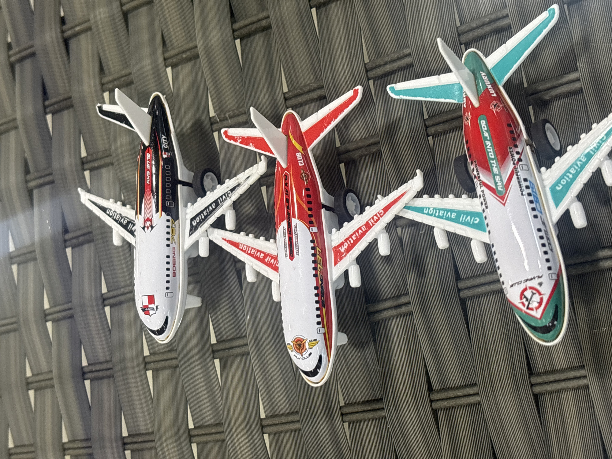 The image shows a top-down view of three toy airplanes lined up on a dark, woven surface, possibly a patio table. The airplanes are positioned vertically in the frame, with the top one slightly to the left and the bottom one slightly to the right. Each plane has distinct livery.
The topmost airplane is predominantly white with a black and red stripe along the fuselage. It has "BOEING 787" printed on it, along with "BLUE SKY" and "CITY." A red and white crest is visible on the nose.
The middle airplane is white with red and yellow accents. It features the text "CIVIL AVIATION," "SPEEDY FLIGHTS," and "CLUB." The "BOEING 737" is also visible.
The bottom airplane is white with teal and red markings. It displays "CIVIL AVIATION," "POWERFUL POWER," and "LUXURY." The text "SOAR INTO THE SKY" is prominently displayed.
The background is a textured, dark gray woven material. The lighting suggests an outdoor setting. No people are visible in the image, and no specific location in Dubai, United Arab Emirates, can be identified.