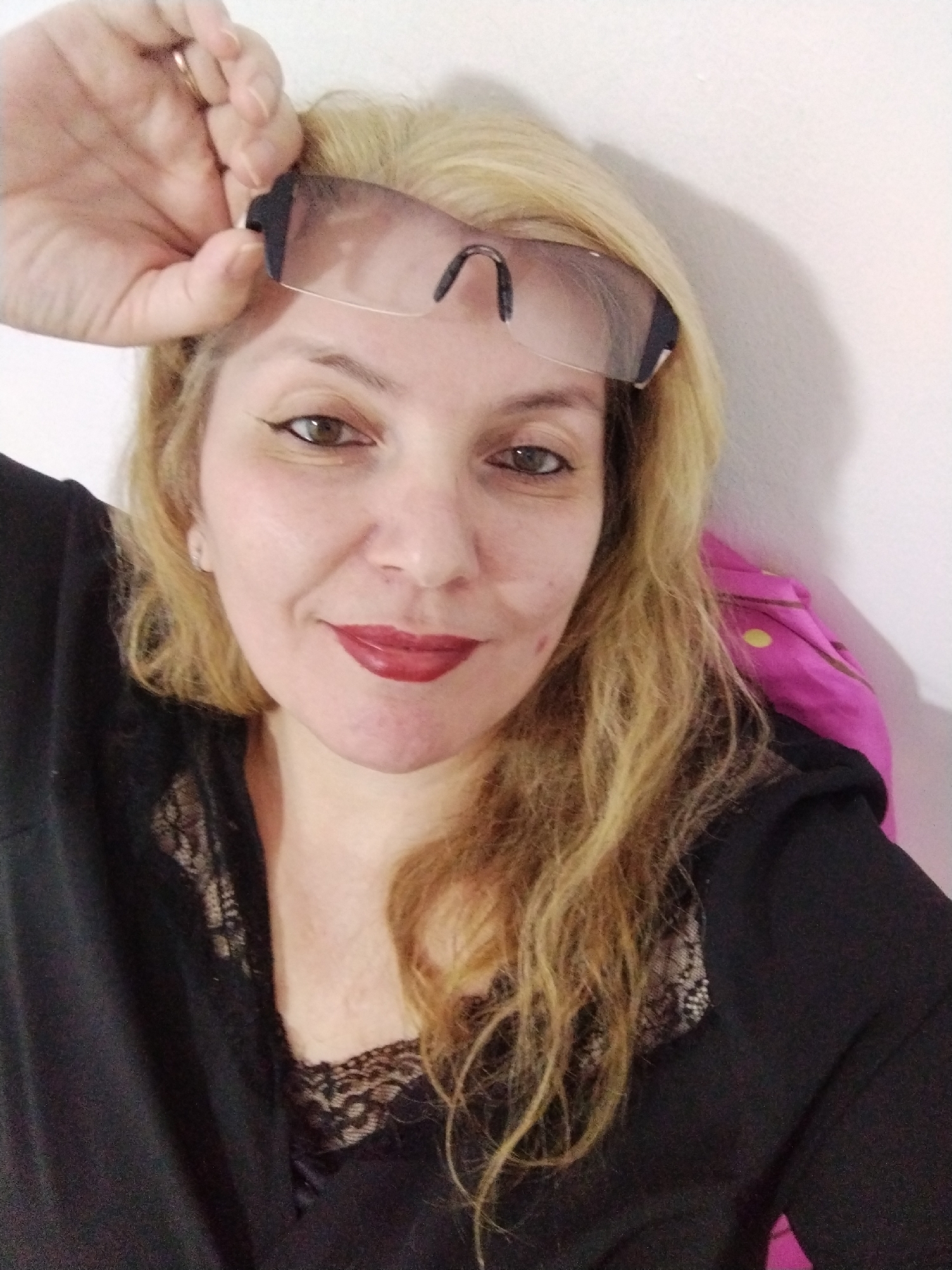 A woman with blonde, wavy hair and a fair complexion is shown from the chest up, looking towards the camera. She is wearing dark clothing with a lace trim and has dark eyeliner and red lipstick. She is holding a pair of clear safety glasses above her forehead with her right hand, and her left hand is visible near her temple. A pink garment with a yellow polka dot pattern is visible on her right shoulder. The background consists of a plain white wall. The image was taken indoors. There is no visible indication of the specific location within Al Mas'oudi, United Arab Emirates.