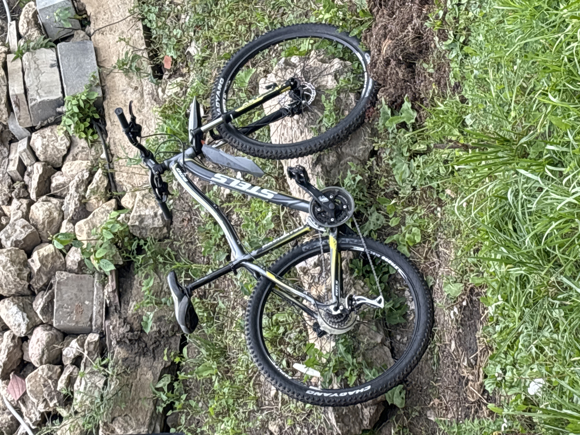 A black and gray Stels mountain bicycle is parked on a grassy incline next to a rough stone wall. The bicycle has a silver fender over the front wheel and a black seat. The front wheel is turned slightly to the left. The background consists of a pile of rough, light-colored stones and some green vegetation. The ground in the foreground is covered in grass and dirt. The scene appears to be outdoors in a natural setting, possibly a park or a wooded area within the city of Zvenigorod, Russia. There are no people visible in the image.