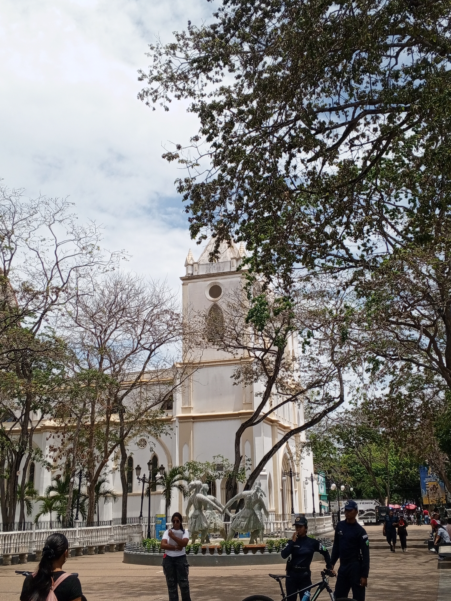 The image captures a vibrant daytime scene in a public square, likely in Porlamar, Venezuela, under a bright, cloudy sky. A large, prominent white church or cathedral dominates the background, featuring a tall, ornate tower topped with a dome, arched windows, and intricate architectural details.
Numerous trees with varied foliage frame the scene; some are full of dense green leaves, while others display intricate networks of bare branches, suggesting different tree types or seasonal changes.
In the midground, in front of the church, a decorative fountain or statue group depicts three stylized figures, seemingly dancing or interacting, surrounded by a white, ornate railing. Streetlights and benches are also visible along the path.
Several people populate the foreground and midground. To the bottom left, the back of a person with dark hair and a pink backpack is seen, possibly near a bicycle. Near the center, a woman in a white t-shirt, dark trousers, and sunglasses stands with her arms crossed. To the right, two uniformed individuals, likely police or security, stand beside a bicycle; the female officer appears to be speaking into a communication device, and the male officer's uniform displays partially visible text, possibly "PRSE".
Further in the background to the right, more people can be seen along a pathway, alongside what appears to be storefronts or market stalls. Visible text on banners includes "FÉNIX" and partially "ARROCOLES" on one, and another partially shows "POLLOS". The overall scene depicts a typical busy day in a communal urban space.