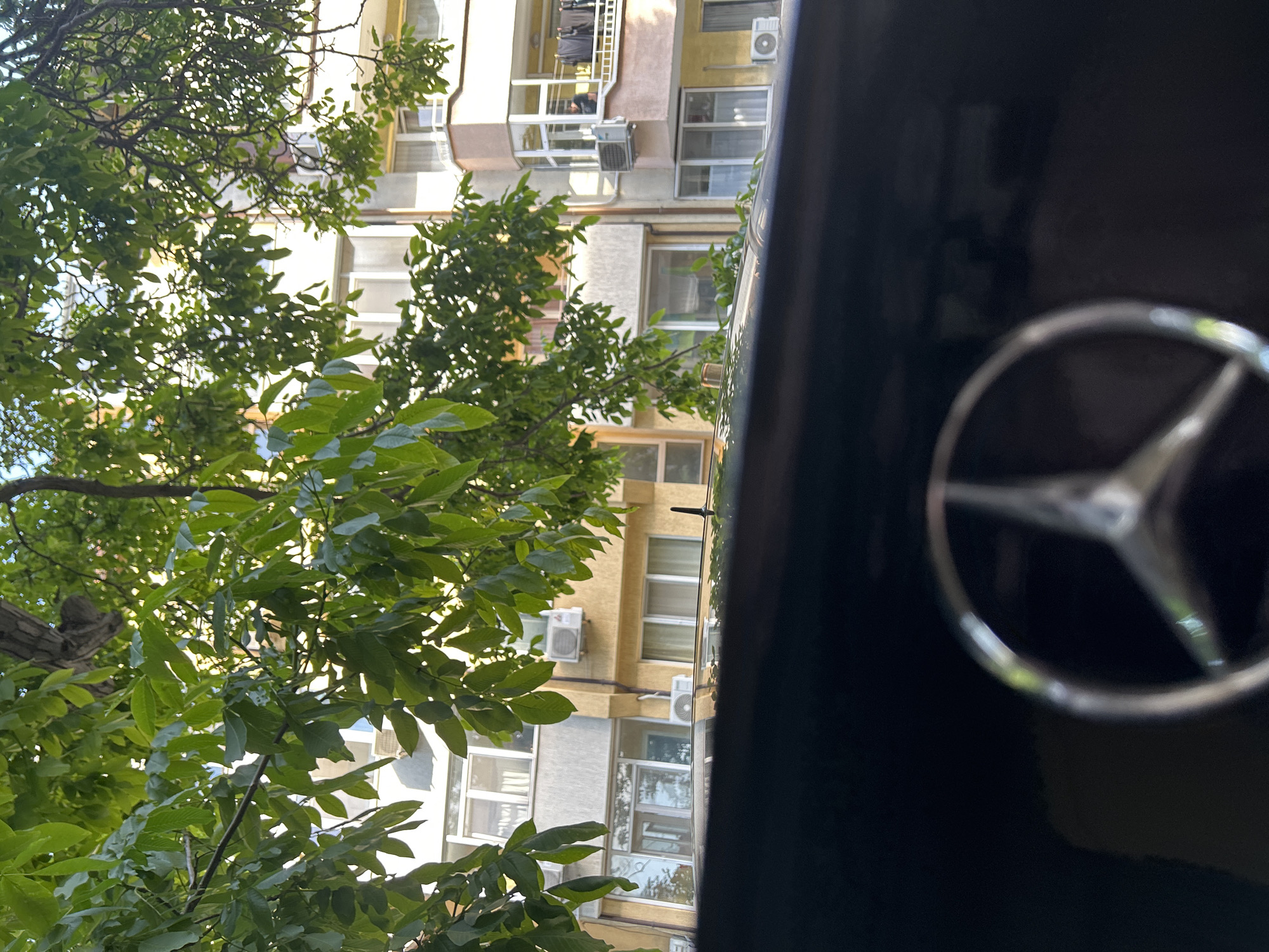The image captures a residential street scene in Varna, Bulgaria, viewed from a slightly elevated perspective, likely from within a parked vehicle. The foreground, occupying the bottom quarter of the frame, shows the dark-colored hood or dashboard of a vehicle, with a blurred, metallic Mercedes-Benz star logo partially visible at the bottom center, and a black car antenna on the right side of the vehicle's roofline.
Beyond the vehicle, multiple multi-story residential buildings are visible, partially obscured by dense green tree foliage that dominates the mid-ground and upper sections of the image. The buildings display varied facades: one on the left appears grey with white-framed windows, while others feature shades of yellow, ochre, and light brown/pink accents on balconies. Numerous external air conditioning units are mounted on the building walls. On a balcony of the rightmost building, dark clothes are hanging to dry. A person, appearing to be an adult male in a dark shirt, is partially visible behind the railing of this balcony, looking left.
The scene is illuminated by natural sunlight, filtering through the vibrant green leaves of the trees, which include a thick trunk visible in the upper left. Small patches of clear sky are discernible through the canopy. The overall environment suggests an urban residential setting during daytime. Specific street or neighborhood identification within Varna is not possible based solely on the visual information provided.