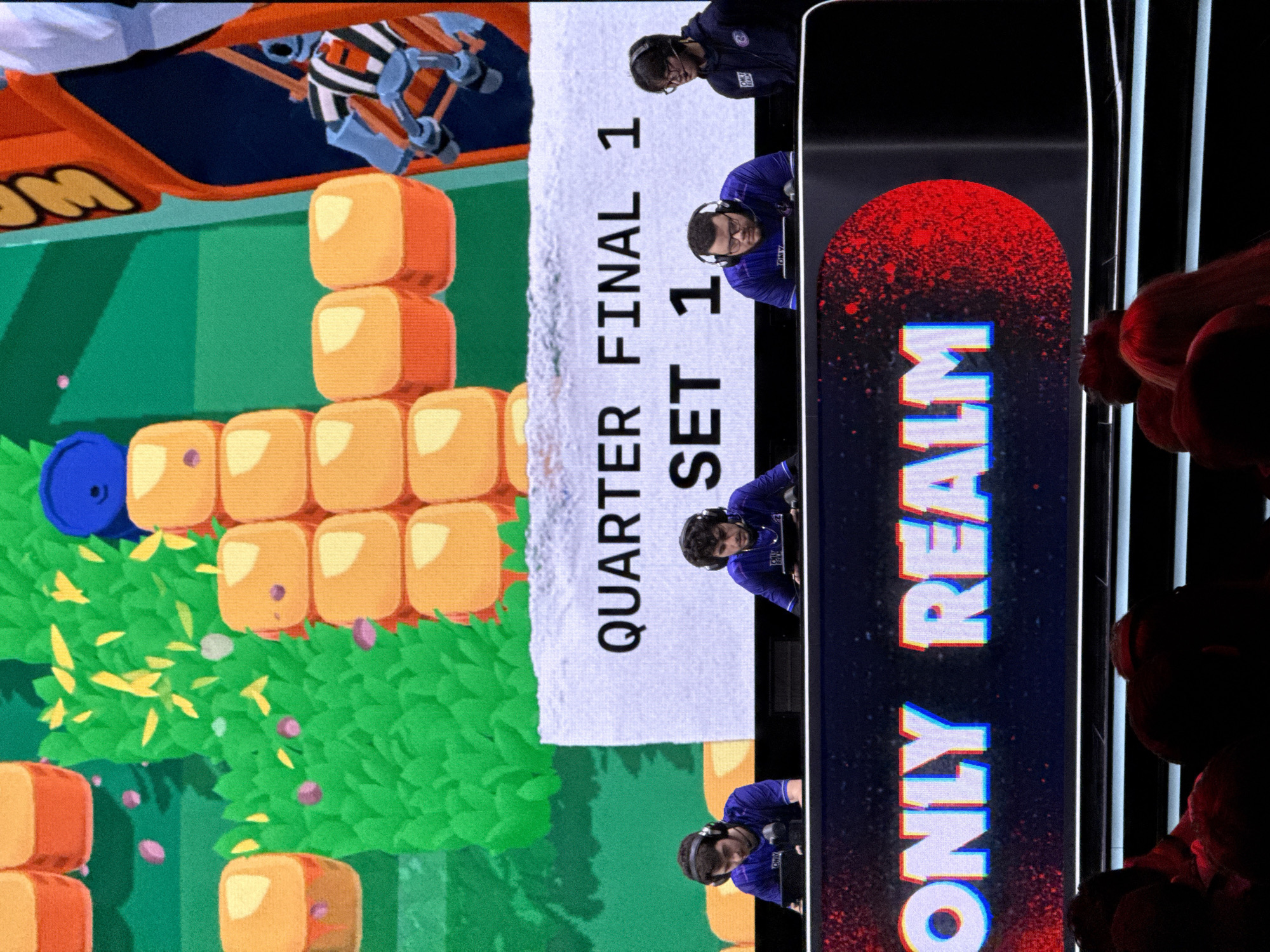 The image depicts a scene from the Quarter Final Set 1 of the "ONLY REALM" competition, held in Berlin, Germany. A digital display shows gameplay footage resembling a blocky, cartoonish environment with orange cubes and green foliage. Overlayed on the gameplay feed is the competition's announcement, as well as several competitors wearing blue shirts, headsets, and facing forward, seemingly focused on the competition. The display background is black with a stylized red splotch pattern, and the words "ONLY REALM" are vertically displayed in white and blue.