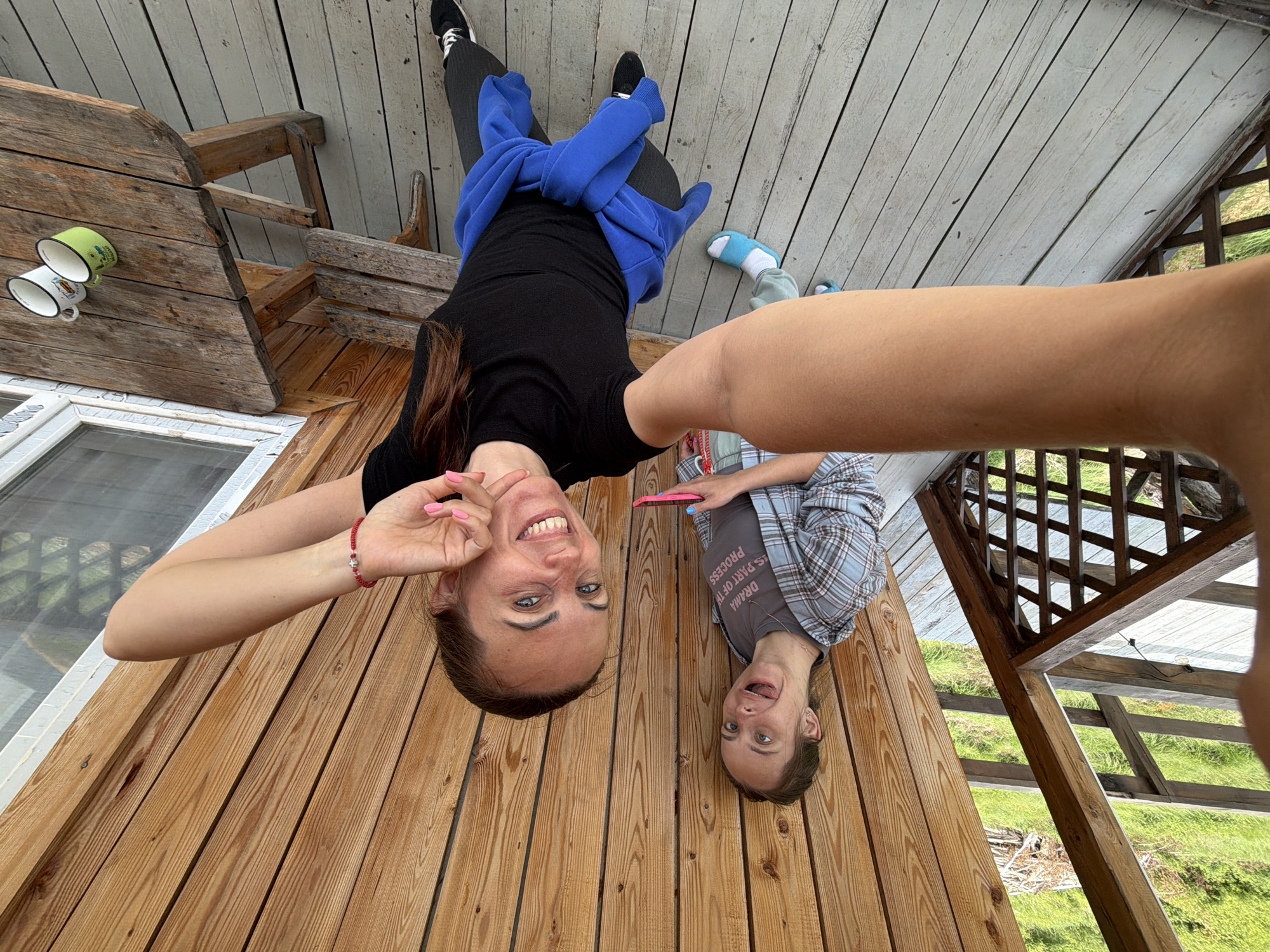 Two women are taking a selfie on a wooden porch overlooking a grassy area with trees. The woman in the foreground is smiling at the camera, wearing a black t-shirt and dark pants, with a blue jacket tied around her waist. The woman in the background is sticking her tongue out at the camera, wearing a grey t-shirt and a plaid shirt. A wooden table with two mugs is visible on the right side of the image. The location is identified as Talakan, Russia.