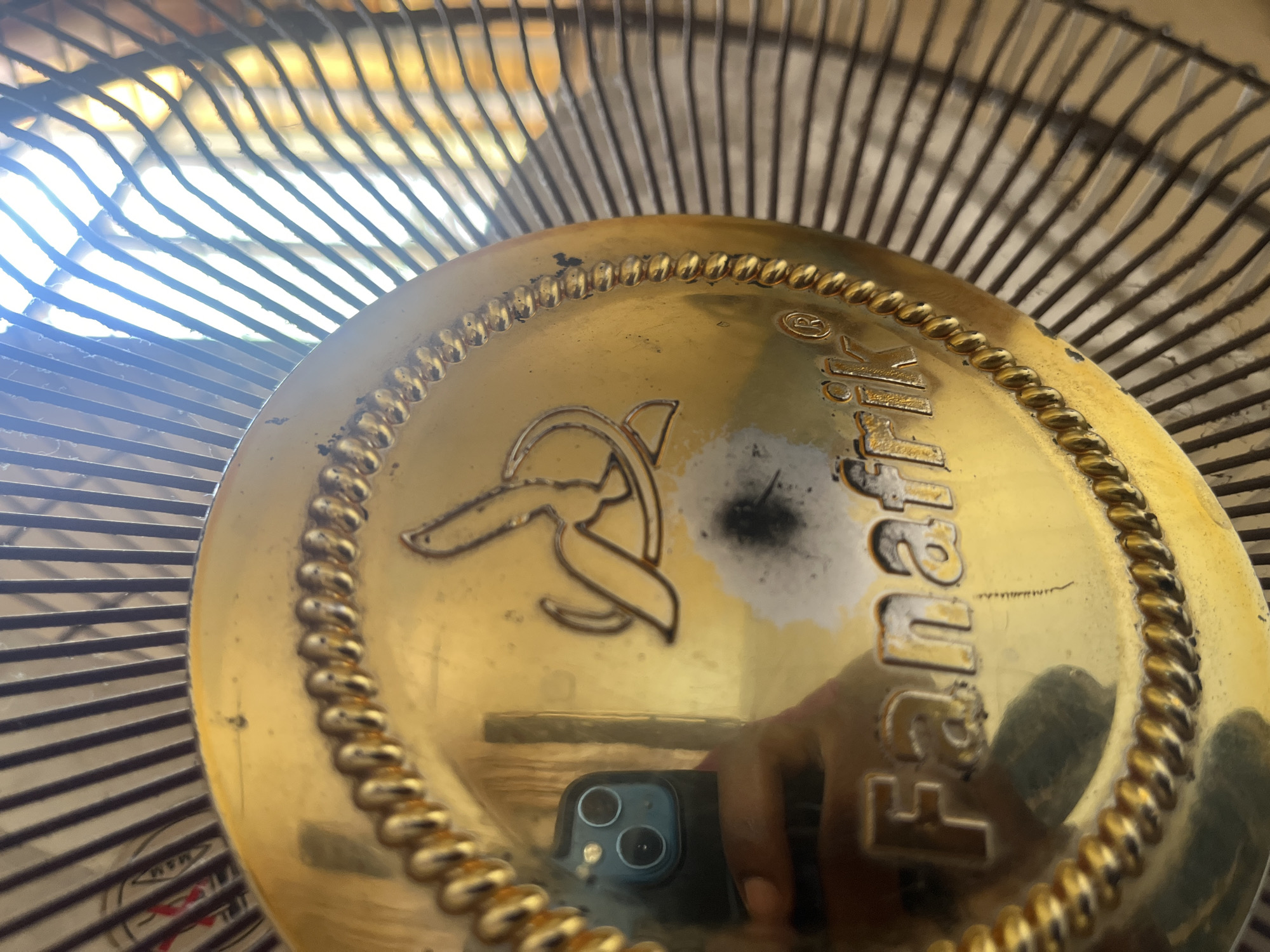 The image shows a close-up of a golden fan grille with the brand name "Fanafrik" embossed on it. A stylized banana logo is also visible. The fan is positioned against a background of metal bars, suggesting it's part of a larger fan structure. Sunlight is streaming through the bars, creating a bright, hazy effect. In the reflection on the golden surface, a smartphone and a hand are visible, indicating that the photo was taken by someone holding a phone. There are some scuff marks and discoloration on the golden surface. The scene appears to be indoors, possibly a room in Jalingo, Nigeria, where such a fan would commonly be found.