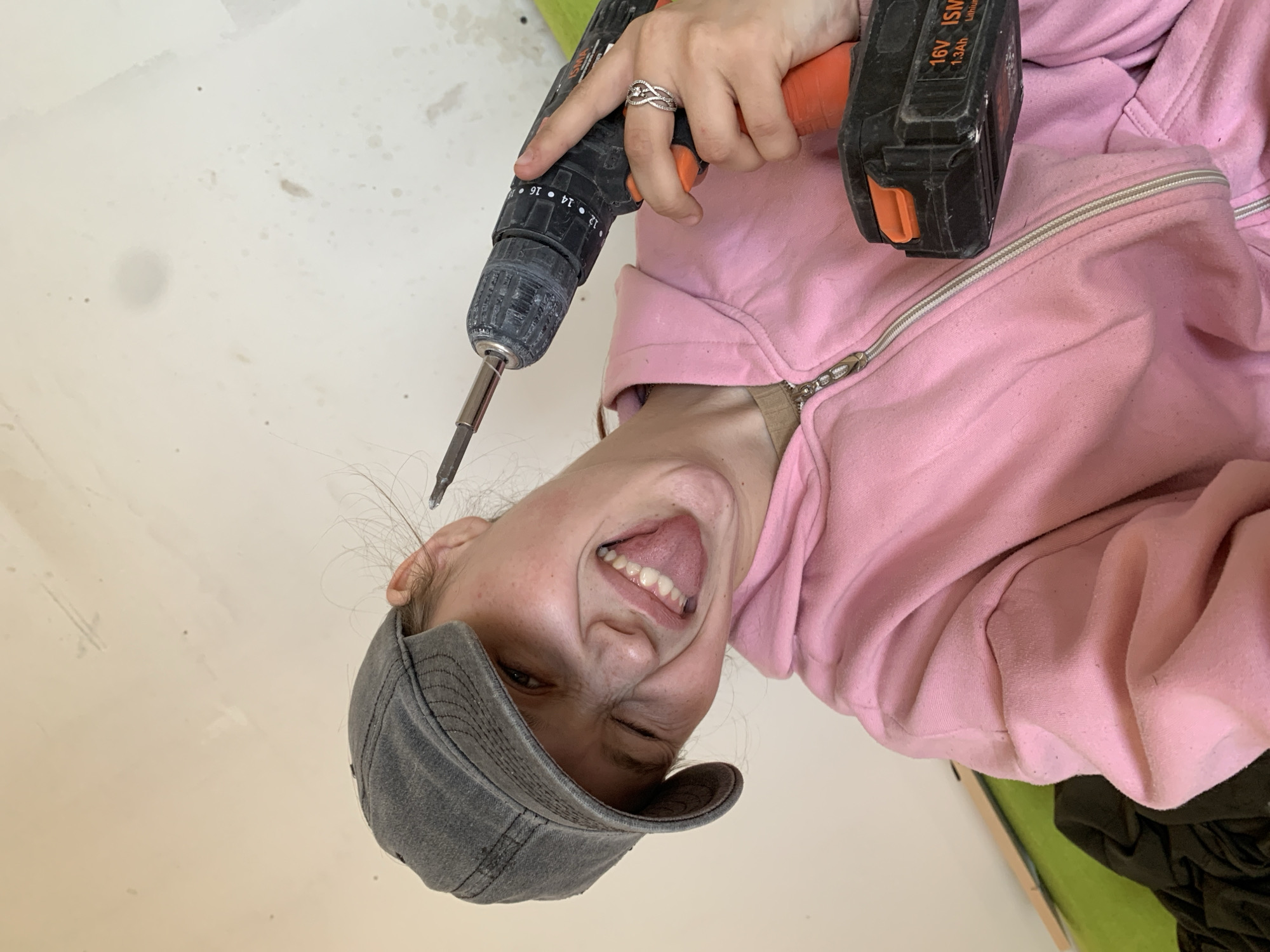 A person wearing a gray baseball cap and a pink hoodie is holding a cordless drill in their right hand with the drill bit pointed towards their face. The person is smiling and sticking their tongue out. The background is a plain white wall. The location cannot be determined from the image.