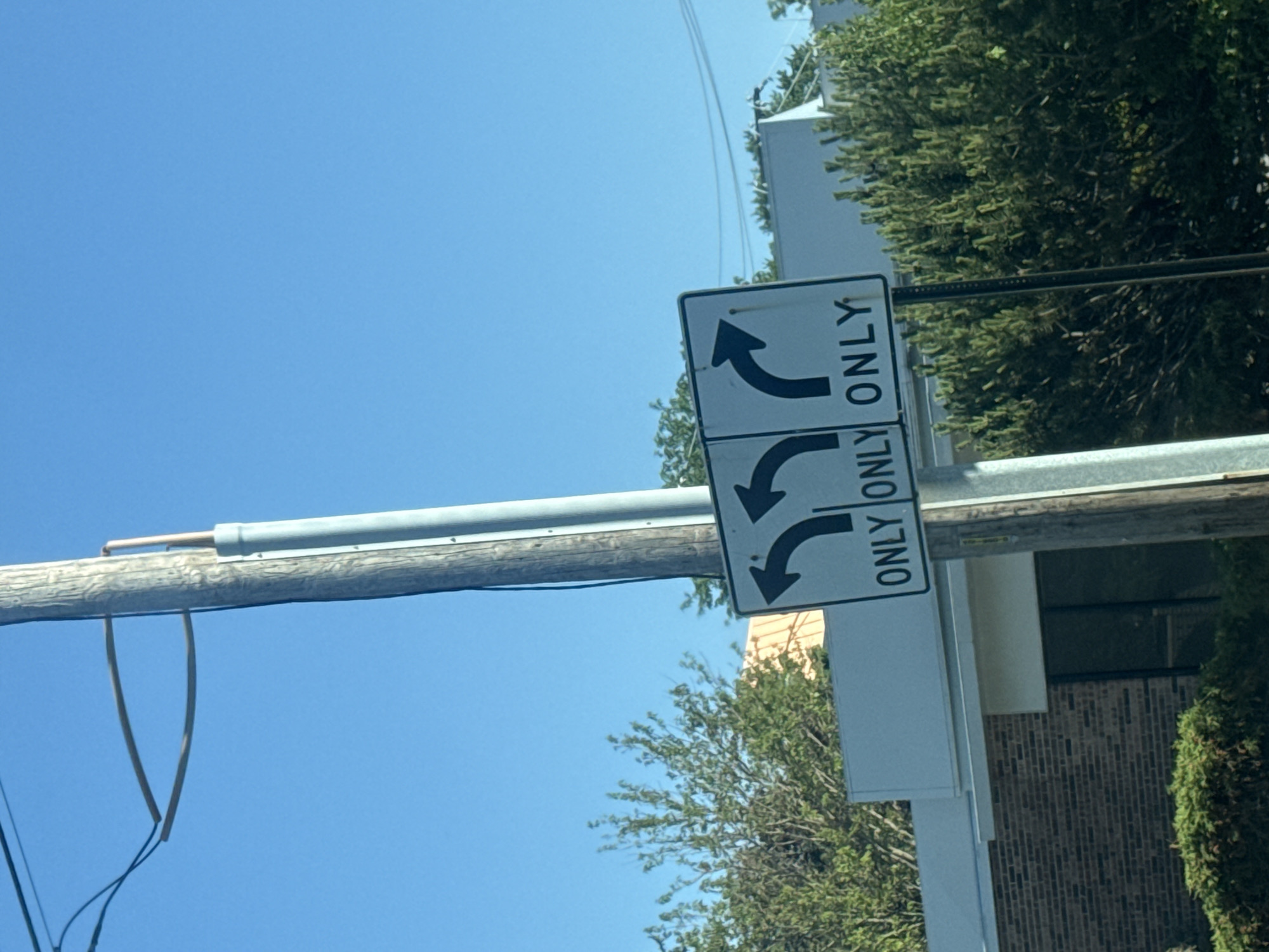 The image shows a street sign indicating mandatory left-turn lanes. The sign is attached to a utility pole. The sign reads "ONLY" next to arrows that curve to the left. The sky is blue and partially visible in the background. Also in the background are green trees and a brick building. While I can identify the country as the United States of America, I cannot specify its exact location as Skokie without additional visual cues or contextual information.