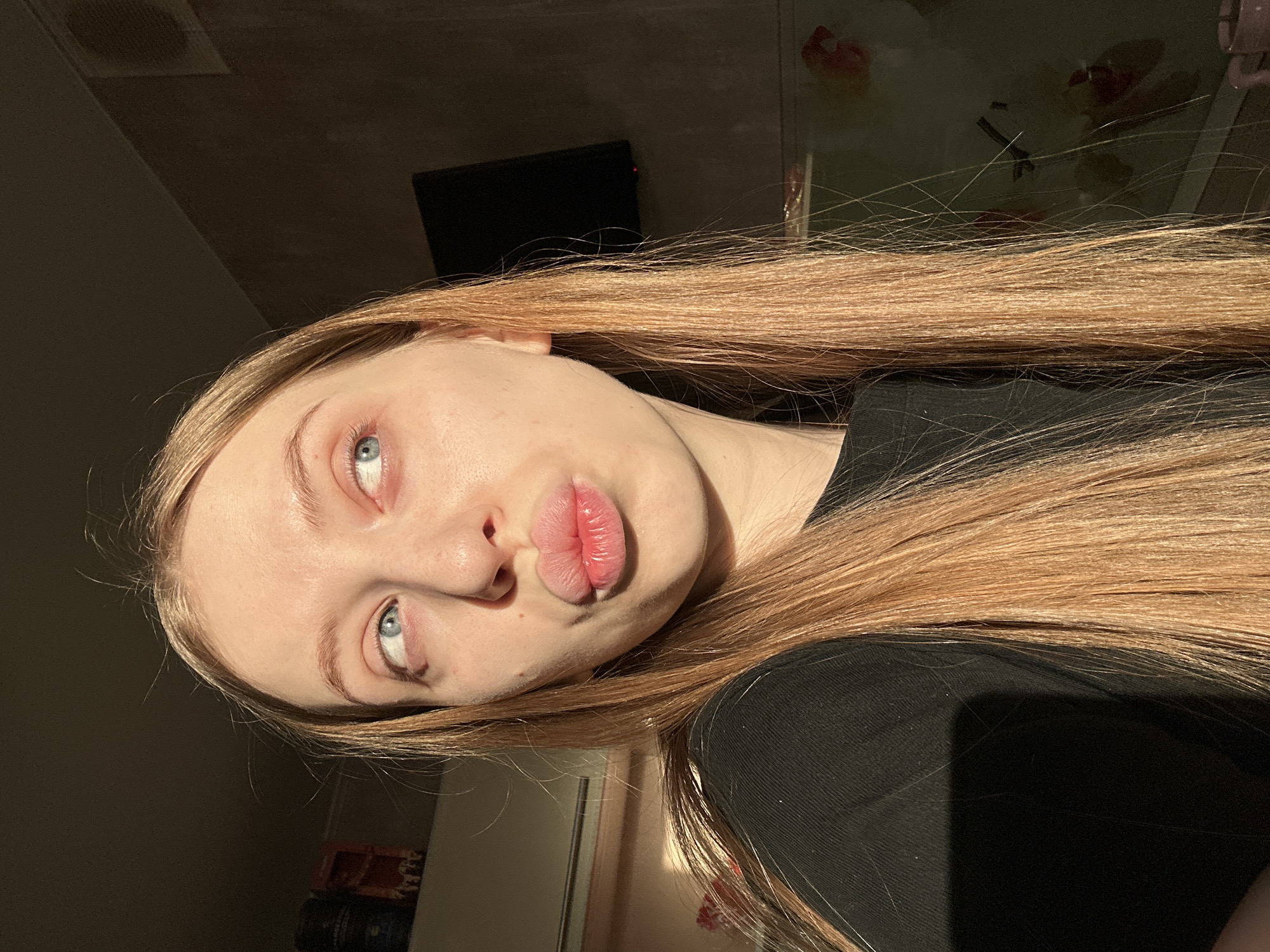 This image depicts a young adult female located indoors in Chaikovsky, Russia. She is shown from the chest up, positioned slightly off-center to the left. The subject has fair skin, long, straight, light brown to auburn hair, and light blue eyes. Her lips are pursed in a "kissing" gesture, and her gaze is directed upwards and to her left (viewer's right). She is wearing a dark, possibly ribbed, short-sleeved top.
The scene is illuminated by strong, warm light originating from the left (viewer's right), casting a distinct shadow across the right side of her face (viewer's left) and illuminating the left side of her face and hair. The background on the left (viewer's right) reveals a white rectangular unit, likely an air conditioner or vent, mounted on a light-colored wall. Above it, on a shelf, are several dark cylindrical containers and other indistinct items. The background on the right (viewer's left) is significantly darker, revealing a dark, rectangular screen (possibly a television or monitor) mounted on a dark wall. Below this screen, faint reflections or translucent decorations with red and orange elements are visible on a surface. A dark vent is also visible in the upper right portion of the background.