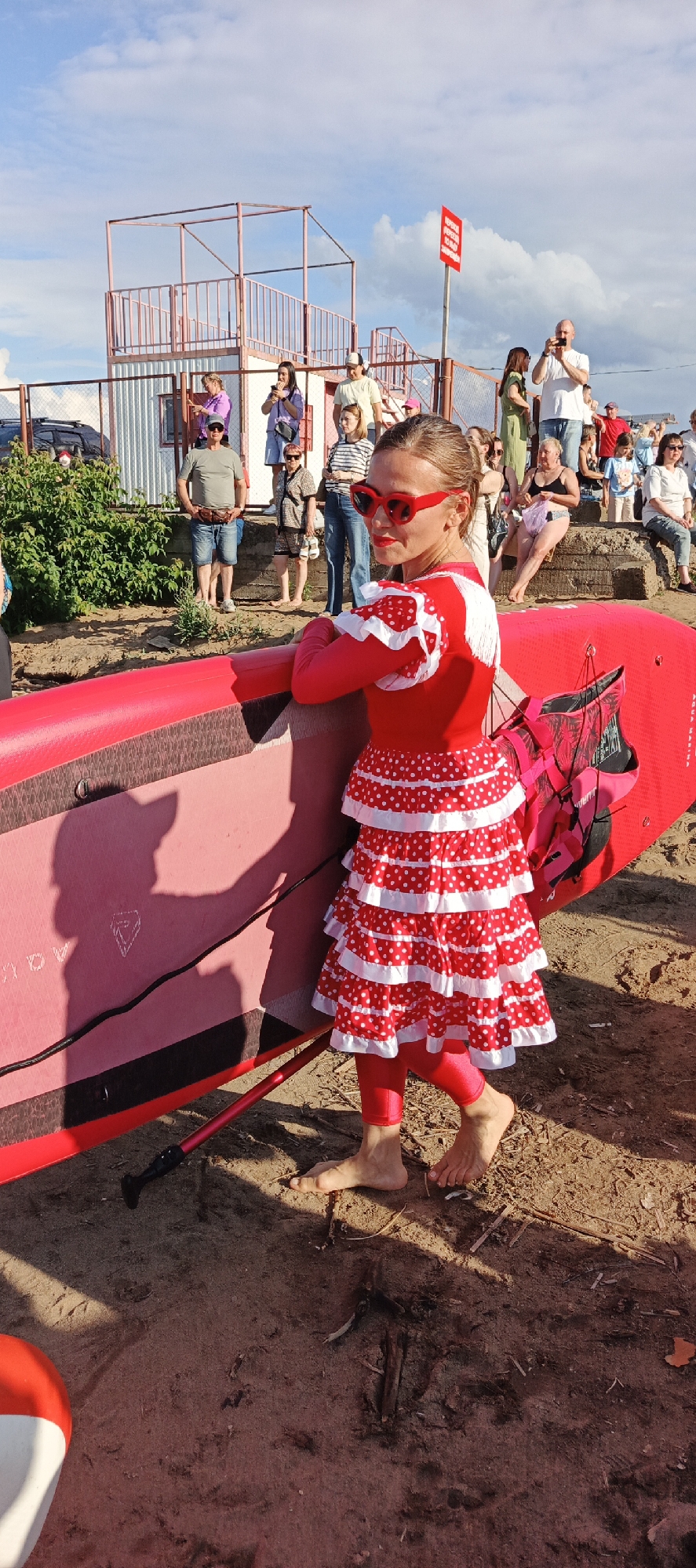 A young girl, dressed in a vibrant red and white polka-dot flamenco-style dress and red leggings, stands on a sandy beach, holding a bright red paddleboard. She wears red sunglasses and has her hair pulled back. Her pose suggests she is looking towards the camera with a slight smile.
In the background, a crowd of people are gathered, some standing, some sitting on concrete steps. They appear to be spectators, with some holding phones to take pictures. Behind them, a small, elevated structure with a metal frame and railings, resembling a spectator stand or platform, is visible. A red and white sign with Russian text is mounted on a pole. The sun is casting long shadows, indicating it is late afternoon or early evening. The sky is blue with scattered clouds. The scene appears to be an outdoor event, possibly a water sports competition or festival, given the presence of the paddleboard and the gathering of spectators. The overall atmosphere seems casual and festive.