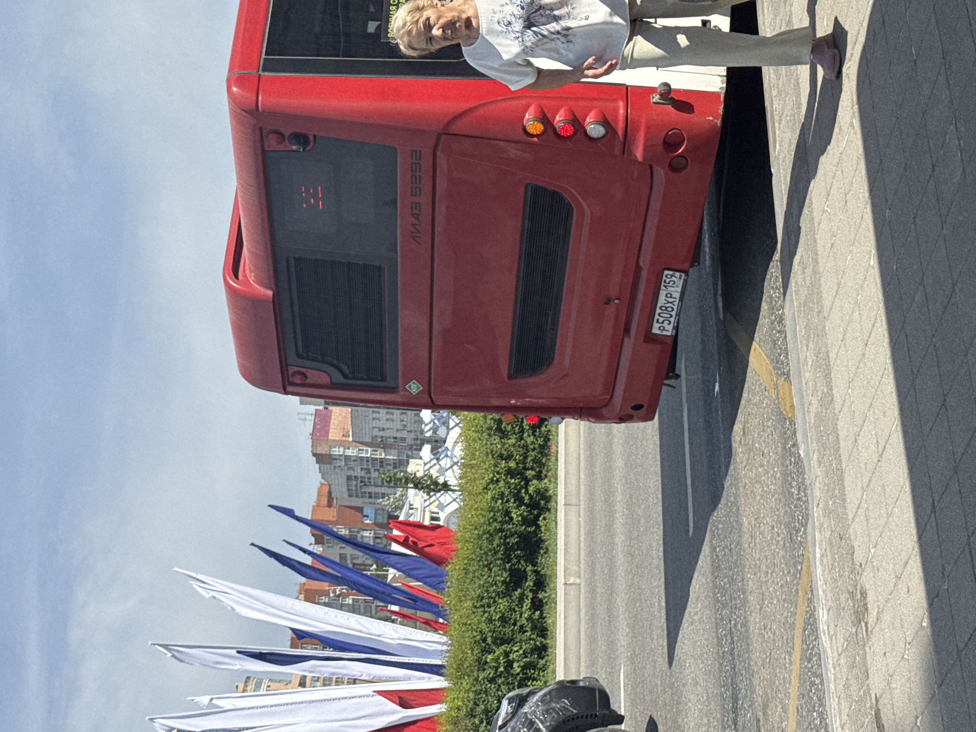 The image depicts a red bus with the Cyrillic letters "ЛИАЗ" and "5292" visible on the rear, along with a license plate reading "P508XP 159." A Caucasian woman is standing near the rear of the bus, wearing a white t-shirt and light-colored pants, and is barefoot. The bus is parked alongside a street with flags in the colors of white, blue, and red lining the road. Green shrubbery separates the flags from the street. A building complex is visible in the background. The setting is in Perm, Russia, as indicated by the "159" region code on the license plate. Part of a black object that might be a vehicle is in the foreground.