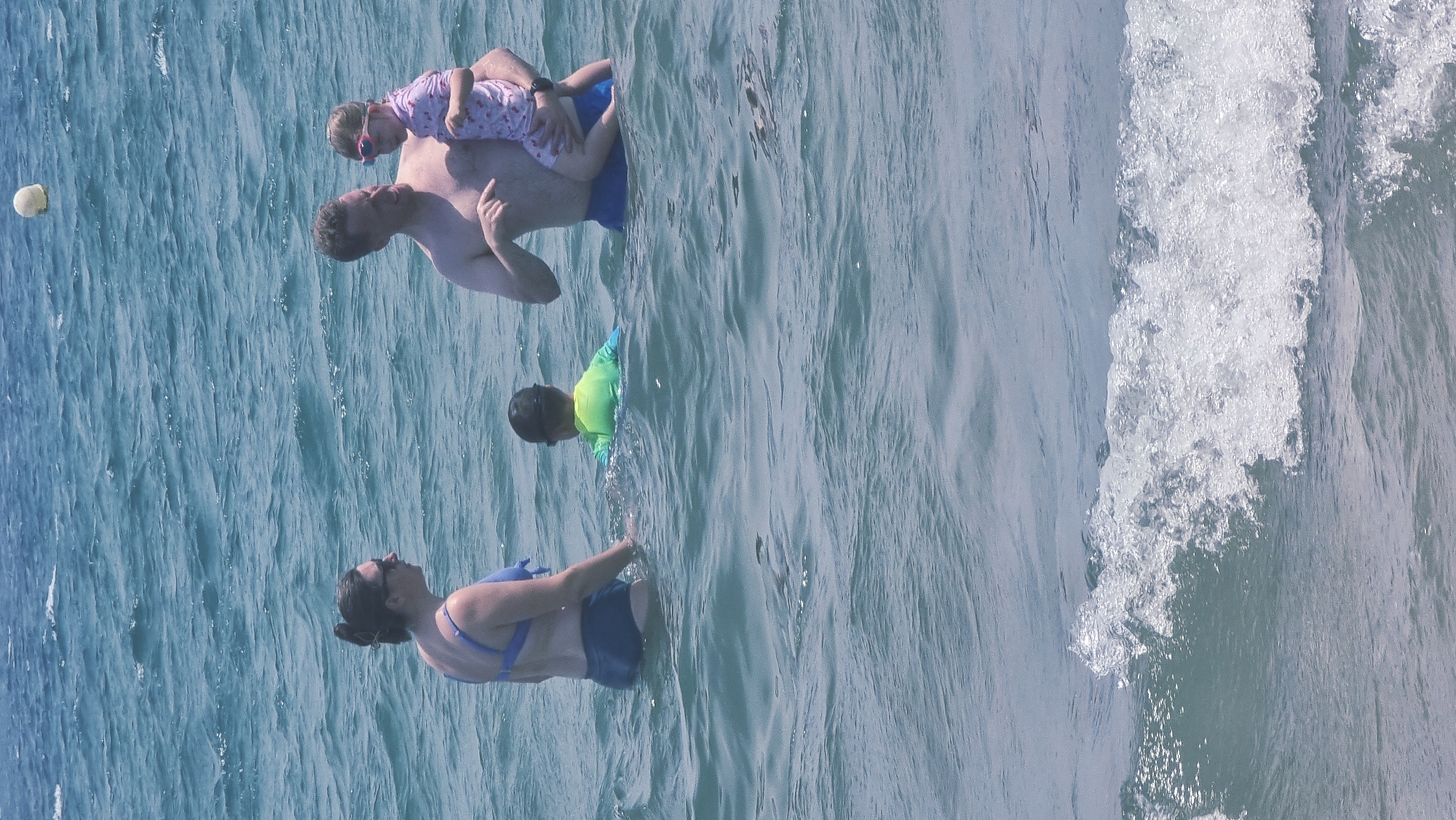 In a sunlit outdoor scene, a family enjoys the ocean. A man, with his back to the viewer, is in the water, holding a young child on his shoulders. The child wears pink pajamas and sunglasses. To the left, a small yellow ball floats on the water's surface. Further into the water, a child in a bright green rash guard is visible. A woman, wearing a blue bikini top and dark bottoms, also in the water, is facing away from the viewer. The water is a clear blue, with gentle waves rolling in. The scene is set at a beach, likely during daytime, given the brightness. The overall mood appears relaxed and joyful, suggesting a family vacation or a pleasant day at the beach. The location is identified as Castelló d'Empúries, Spain. No text is visible in the image.