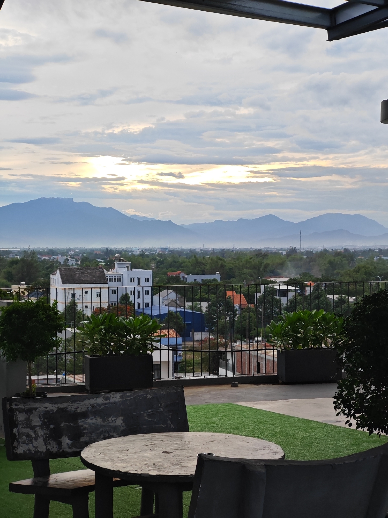 The image depicts a rooftop view of a city, likely Đà Nẵng, Vietnam, with mountains in the background. The foreground features an outdoor seating area with a round concrete table and a concrete bench. Artificial grass is visible on the rooftop, alongside potted plants in rectangular planters. A black metal railing separates the rooftop from the buildings and greenery below. In the mid-ground, there are various buildings, including a white structure with multiple windows and a dome-like feature on its roof, surrounded by trees and other structures. The background consists of a hazy landscape with lush green trees and a series of layered mountains under a cloudy sky with sunlight breaking through. The overall scene suggests a tranquil, possibly urban, environment with a natural backdrop, likely captured during dawn or dusk due to the lighting conditions.