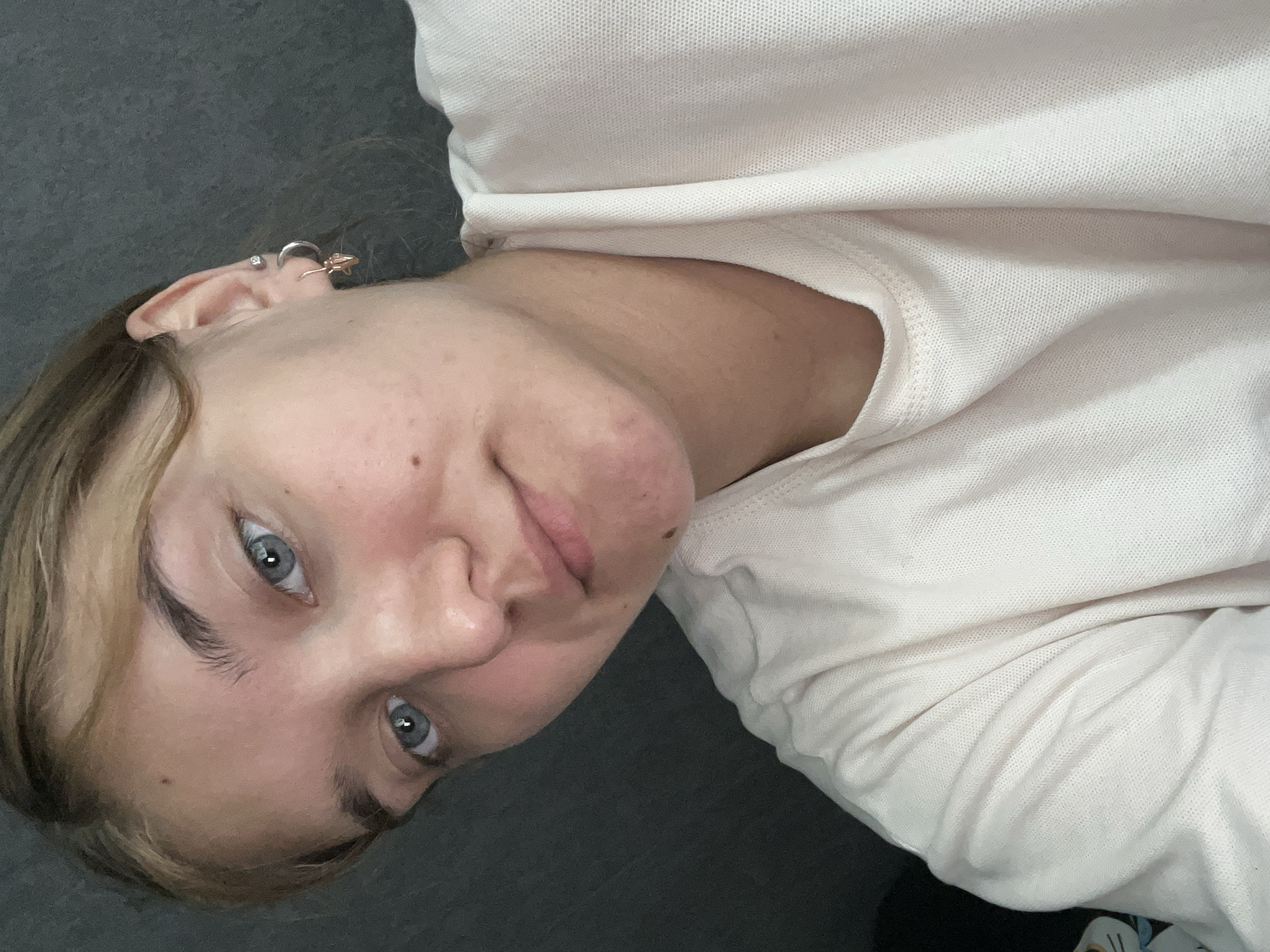 A close-up, waist-up shot features a young Caucasian female with fair skin and blonde hair tied back. She is wearing a light-colored t-shirt. Her eyes are blue and looking towards the camera. She has a small mole on her chin. The background is a plain, dark gray surface. The image was taken in Chaikovsky, Russia.
