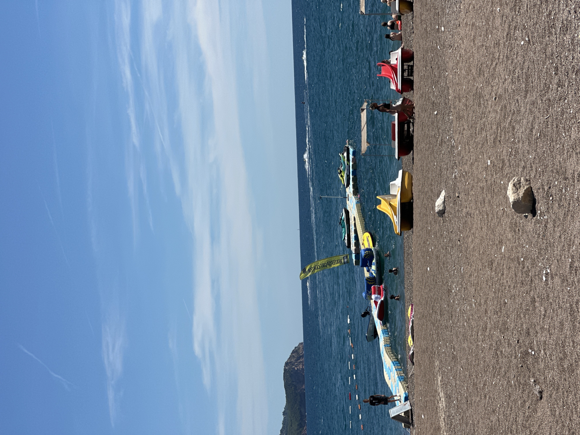 A wide shot captures a sunny day at the beach in Boreti, Montenegro. The foreground shows a dark pebble beach with several large rocks scattered across it. In the middle ground, a line of colorful paddle boats with canopies are parked on the sand, with some people visible sitting on them. To the left, a man stands on a floating pontoon walkway that extends into the turquoise water. Several inflatable toys and smaller boats are also attached to the pontoon. In the water, a few people are swimming and enjoying the sea. Further out, a long yellow flag with text is planted on a floating platform, and several jet skis are being ridden by people, leaving white wakes on the water. In the background, a hilly, tree-covered island is visible in the distance under a clear blue sky with a few wispy clouds and contrails.