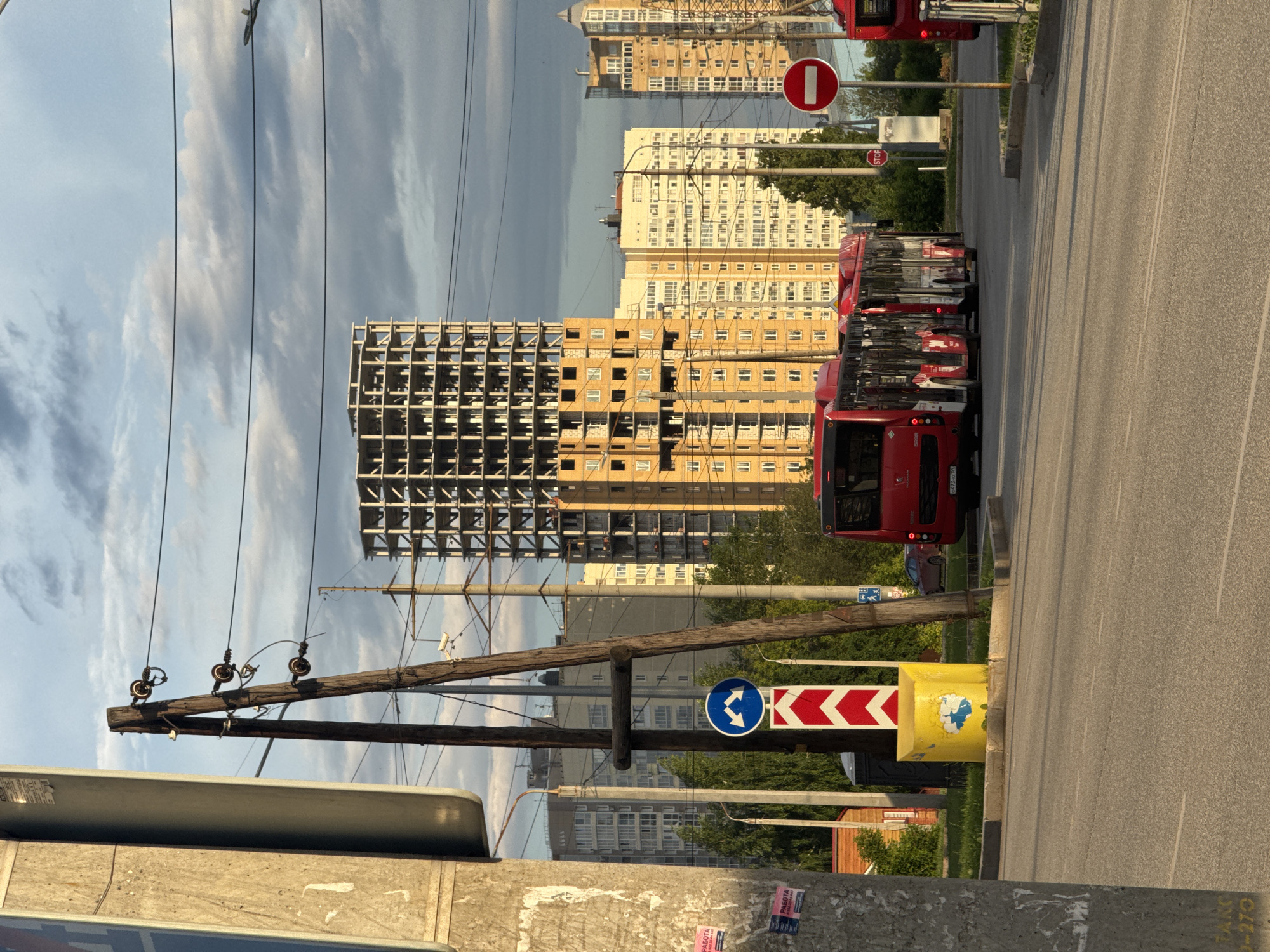 The image depicts a street scene in Perm, Russia, with a focus on a construction site and public transportation. In the foreground, a paved road is visible, with white lane markings. To the left, a large concrete pole with peeling posters stands prominently. Further into the scene, a wooden utility pole with electrical insulators and wires crosses the frame.
A red public bus is positioned on the right side of the road, its side facing the camera, suggesting it is stopped or in motion. Several other red buses are visible behind it, partially obscured. A traffic sign with two arrows indicating left and right turns is present, mounted on a pole with a red and white chevron marker below it. A yellow cylindrical object with a blue drawing of a cloud and a figure is situated near the traffic sign.
In the background, several multi-story buildings are under construction, characterized by their bare concrete frames and exposed rebar. One building is particularly tall and unfinished. Other completed buildings, likely residential apartments, are also visible, featuring yellow and cream facades with numerous windows. The sky above is partly cloudy, with the setting or rising sun casting a warm light on the scene. Trees and greenery can be seen interspersed between the buildings. A red stop sign is visible further down the road. The overall impression is of an urban environment undergoing development, with a clear presence of infrastructure and everyday city life.
