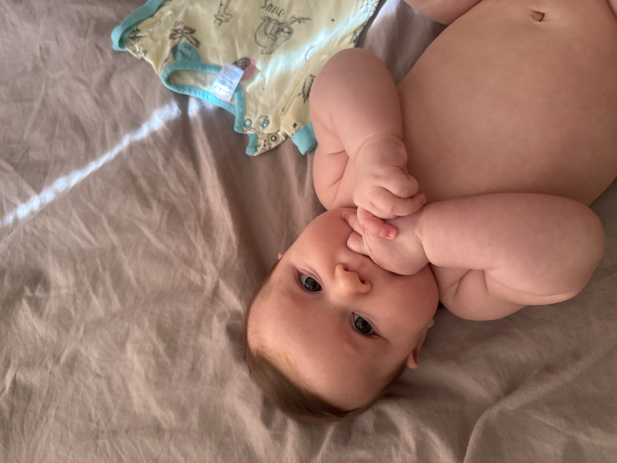 A baby lies on a gray sheet with a yellow and blue patterned onesie nearby. The baby is on its back, with hands raised, and one is in its mouth. The image was taken indoors, during the day. The baby is Caucasian, with fair skin and dark eyes. There is nothing in the image to indicate that it was taken in the city of Chaikovsky, Russia.