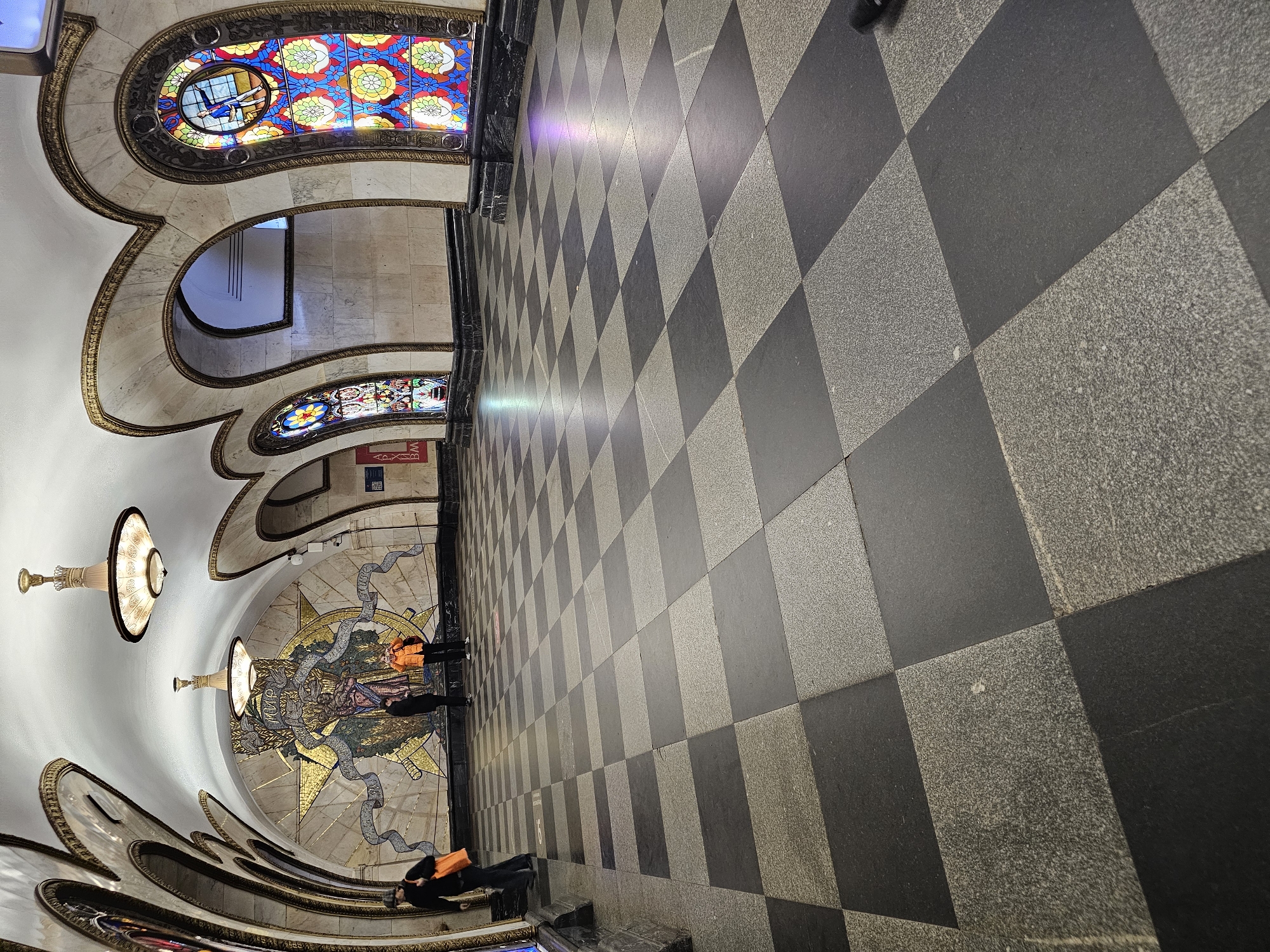 This media file presents an ornate, grand interior space, likely a metro station in Moscow, captured from a perspective rotated 90 degrees clockwise, looking down a long hall or passage. The architectural style is opulent, featuring high, arched ceilings and walls rendered in white or light-colored stone, elaborately trimmed with intricate golden or bronze decorative moldings.
Several large, colorful stained-glass windows are embedded within these arched structures. They display vibrant hues of red, blue, yellow, and green, with detailed floral and geometric patterns. One prominent stained-glass panel clearly depicts a figure, appearing to be an aviator or pilot. Multiple classical-style, dome-shaped chandeliers with decorative bases are suspended from the ceiling, illuminating the space.
On the left side of the image, set into a wall or ceiling arch, is a large, circular mosaic. It features a central figure, possibly a woman or goddess, with flowing ribbons, surrounded by a radiating golden starburst pattern. Near this mosaic, two individuals are standing, one wearing a bright orange jacket. Further in the foreground, another person with an orange top is seated or leaning on a step or ledge.
The floor is a distinctive checkerboard pattern, composed of alternating squares of light grey and dark grey granite or stone tiles, stretching into the distance. A red sign with white Russian text "ВЫХОД" (VYKHOD), meaning "EXIT," is visible beneath one of the stained-glass windows. The number "16" is faintly visible on the checkered floor on the left side, near the seated person. The scene conveys a sense of historical grandeur and artistic detail typical of the Moscow Metro.