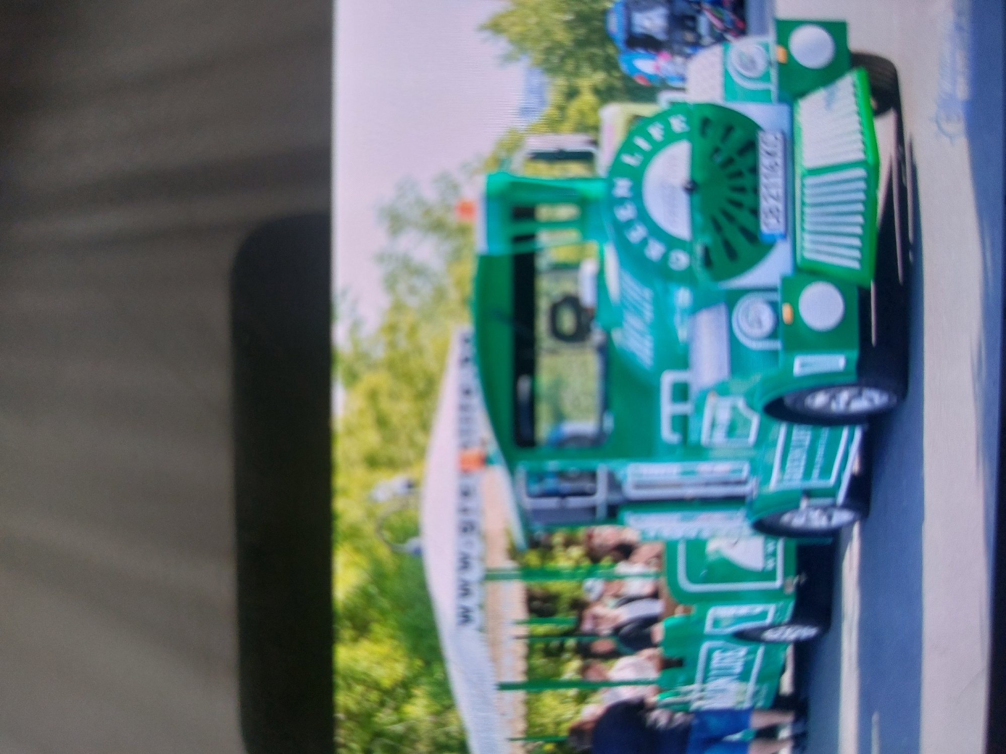 A green sightseeing train, labeled "GREEN LIFE," is shown driving down a road. People are seated in the train's open carriages. The train's front grill displays the license plate "CB 2116 XC". Trees are visible in the background. The image suggests the train is in operation, offering a tour to passengers. Location: likely Tsarevo, Bulgaria, given the vehicle's presence and the visual style.