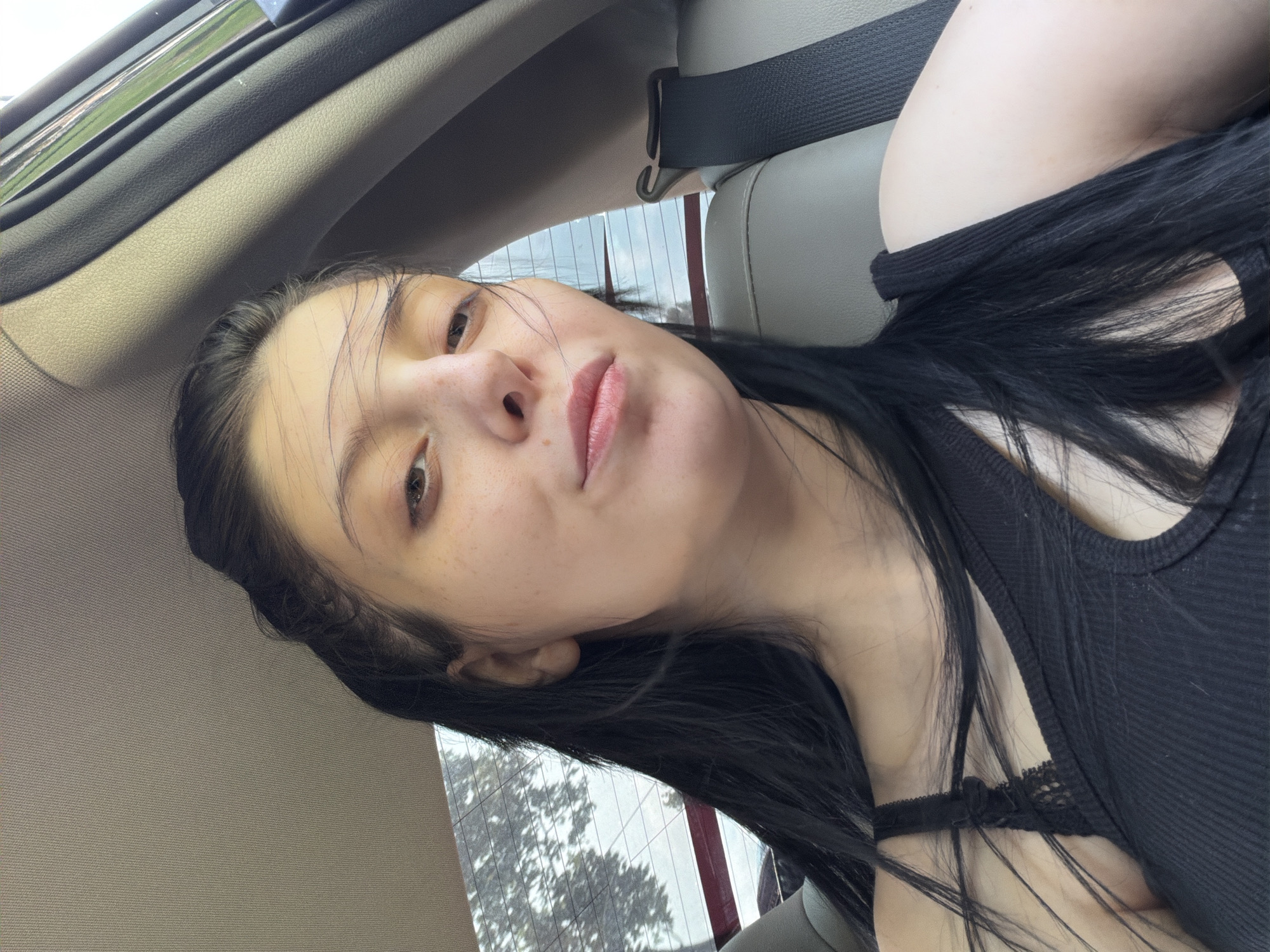A young woman with long, dark hair and fair skin is sitting in the passenger seat of a car. Her head is tilted to her left, and she is looking at the camera. She has light brown eyes and freckles on her cheeks and nose. She is wearing a black, ribbed tank top with lace detailing on the straps. The background shows the interior of the car, including a gray leather seat, a black seatbelt, and the car's ceiling. Through the window, trees and the sky are visible. The image appears to be a selfie taken by the woman.