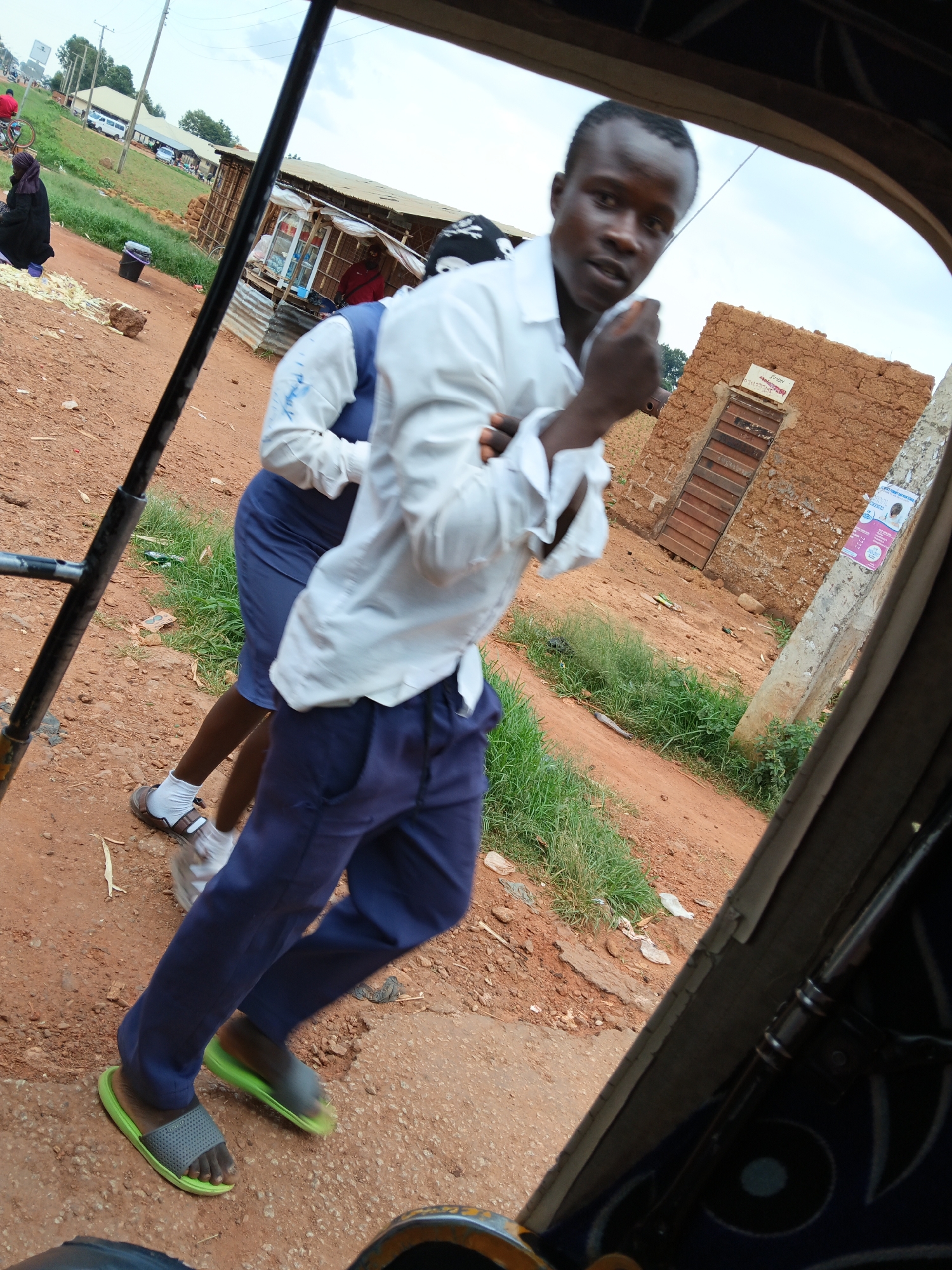 This media file captures a candid scene on a dirt road, likely in a rural or developing area. The foreground is dominated by the interior of a vehicle, with dark framing suggesting a tuk-tuk or similar open-air transport. The primary subjects are two individuals. One is a young man in the foreground, looking towards the camera with a surprised or engaged expression, his left hand raised in a gesture of emphasis. He is wearing a white collared shirt, unbuttoned at the top, and dark blue trousers. His feet are shod in mismatched open-toed sandals, one gray and one lime green. Behind him, another person, likely female given the attire, is visible from the waist down, wearing a dark blue dress and white socks with sandals. Their movement suggests they are walking or getting out of the vehicle.
The background reveals a roadside setting. To the left, a woman in a dark head covering is partially visible, crouched near a pile of yellow items, possibly food produce. Beyond her, a row of small, simple structures with corrugated metal roofs line the road, appearing to be market stalls or small shops. A white van and a bicycle are also visible in the distance, suggesting ongoing activity. To the right, a building constructed from mud bricks with a wooden door stands prominently. A concrete pole with a faded poster attached stands in front of it. The ground is a dusty reddish-brown dirt, with patches of green grass and scattered debris.
The lighting suggests it is daytime, with a bright, overcast sky. The overall impression is one of everyday life unfolding in a busy, yet humble, environment. There is no explicit text in the image that is clearly legible, apart from what appears to be on the poster on the pole, which is too indistinct to read.