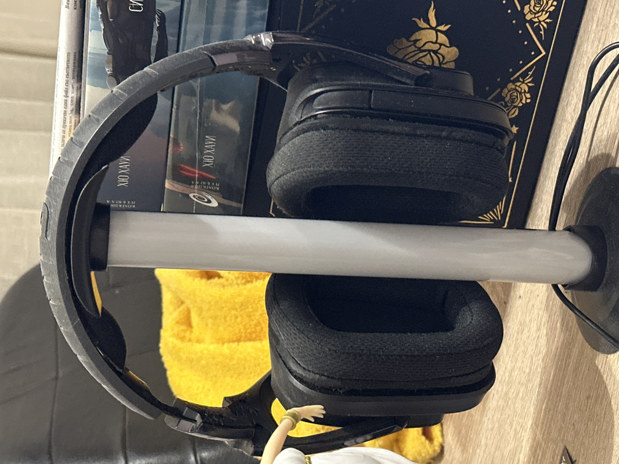 A black over-ear headset is depicted resting on a light grey cylindrical stand with a black circular base, positioned on a light brown wooden surface. The headset features black fabric-covered earcups, exhibiting visible texture and slight compression, and a black headband with an underlying comfort strap. A black electrical cable extends from the stand's base onto the wooden surface. To the immediate left of the headset, the partial view of a white figurine with a gold armband is visible, its right hand positioned near the left earcup. Behind the figurine and the left side of the headset, a crumpled yellow fabric object is present, against a darker, possibly textured, background. Behind the headset stand and the right earcup, several books are stacked horizontally. The visible book spines display Cyrillic text, including "ХЮ ХАУИ" (Hugh Howey) on multiple volumes. The lowermost visible book has a dark cover adorned with gold floral patterns, including roses. The scene is located in the city of Unknown, Unknown.